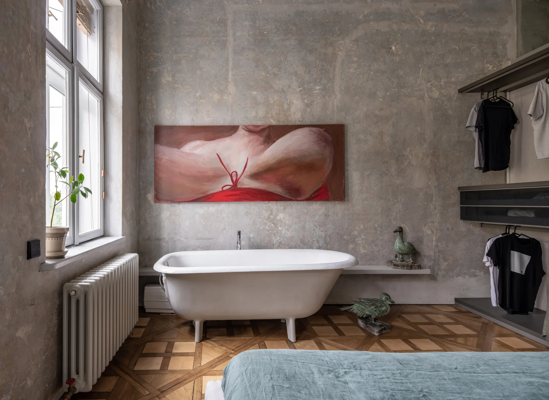 A white freestanding bathtub sits on parquet floors against a textured grey plaster wall decorated with a large horizontal figure painting.