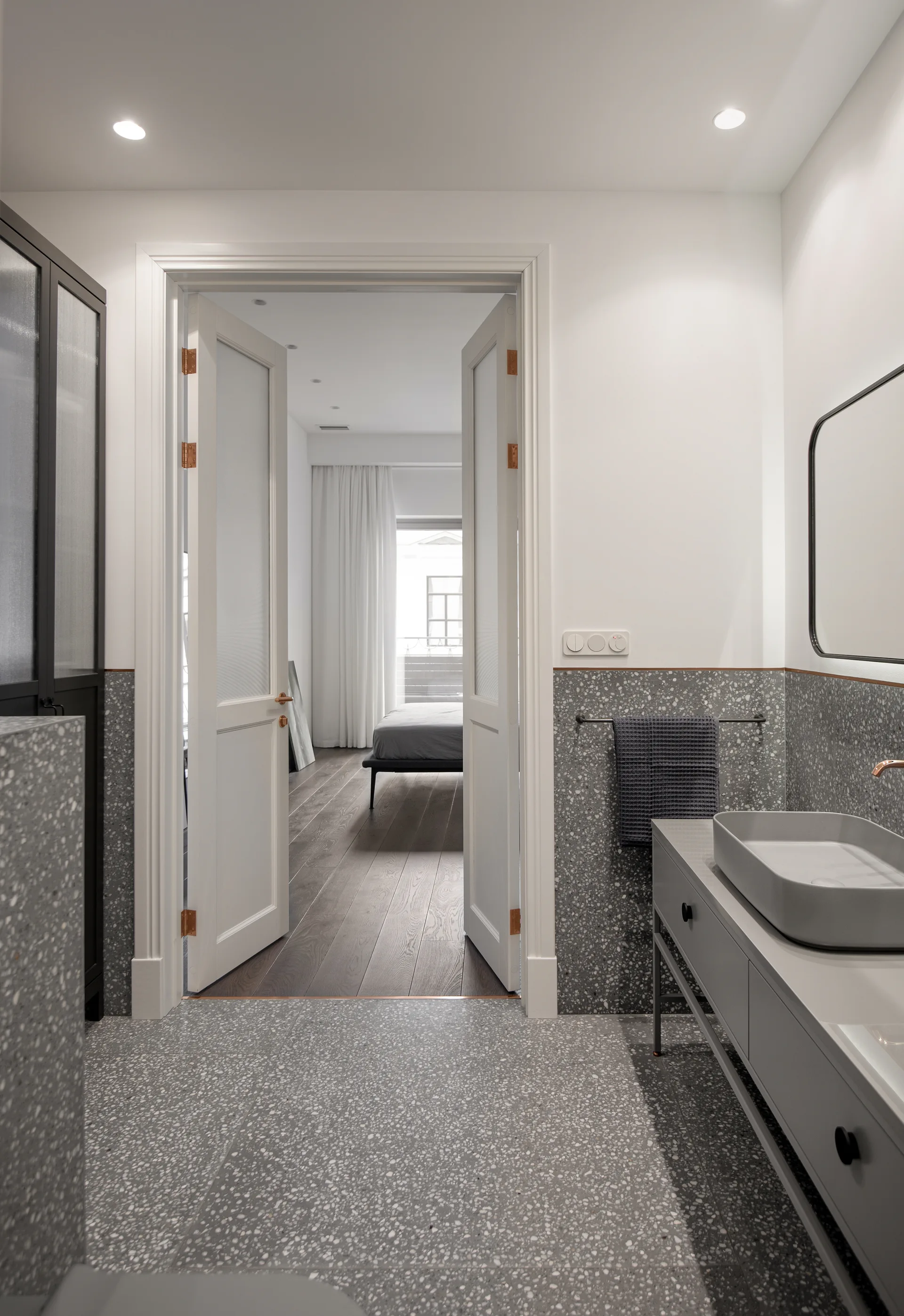 A modern grey terrazzo bathroom features a grey vanity with a vessel sink and open white double doors leading into an adjacent bedroom.