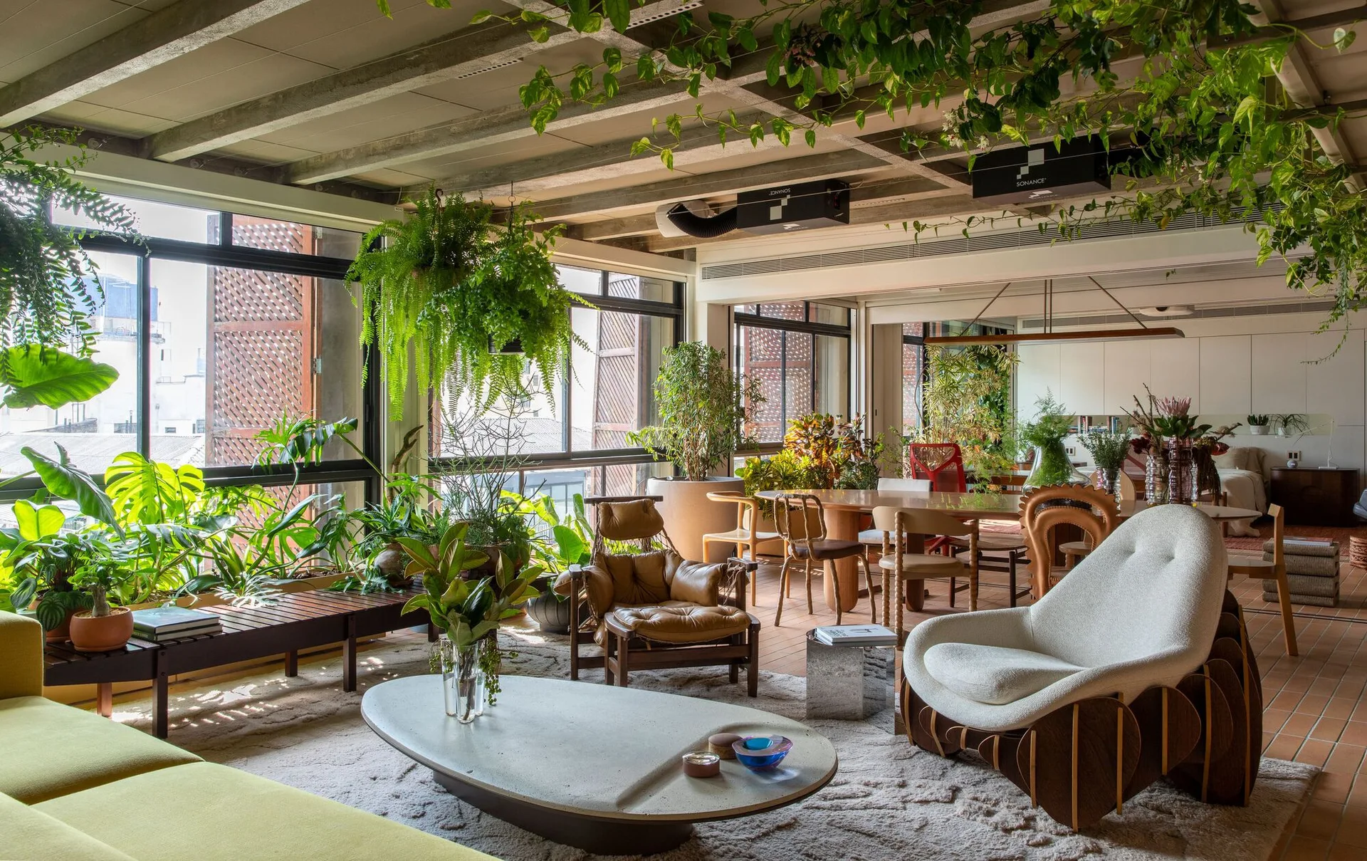 An open-concept living area with concrete ceiling beams, abundant potted plants, a low oval coffee table, and terracotta tile flooring.