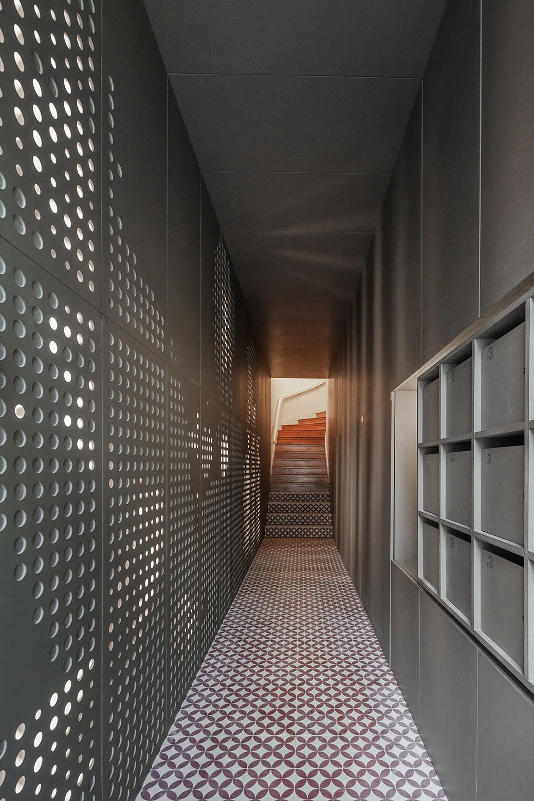 A narrow charcoal gray hallway featuring a backlit perforated metal wall, patterned floor tiles, and built-in mailboxes leading to a wooden staircase.
