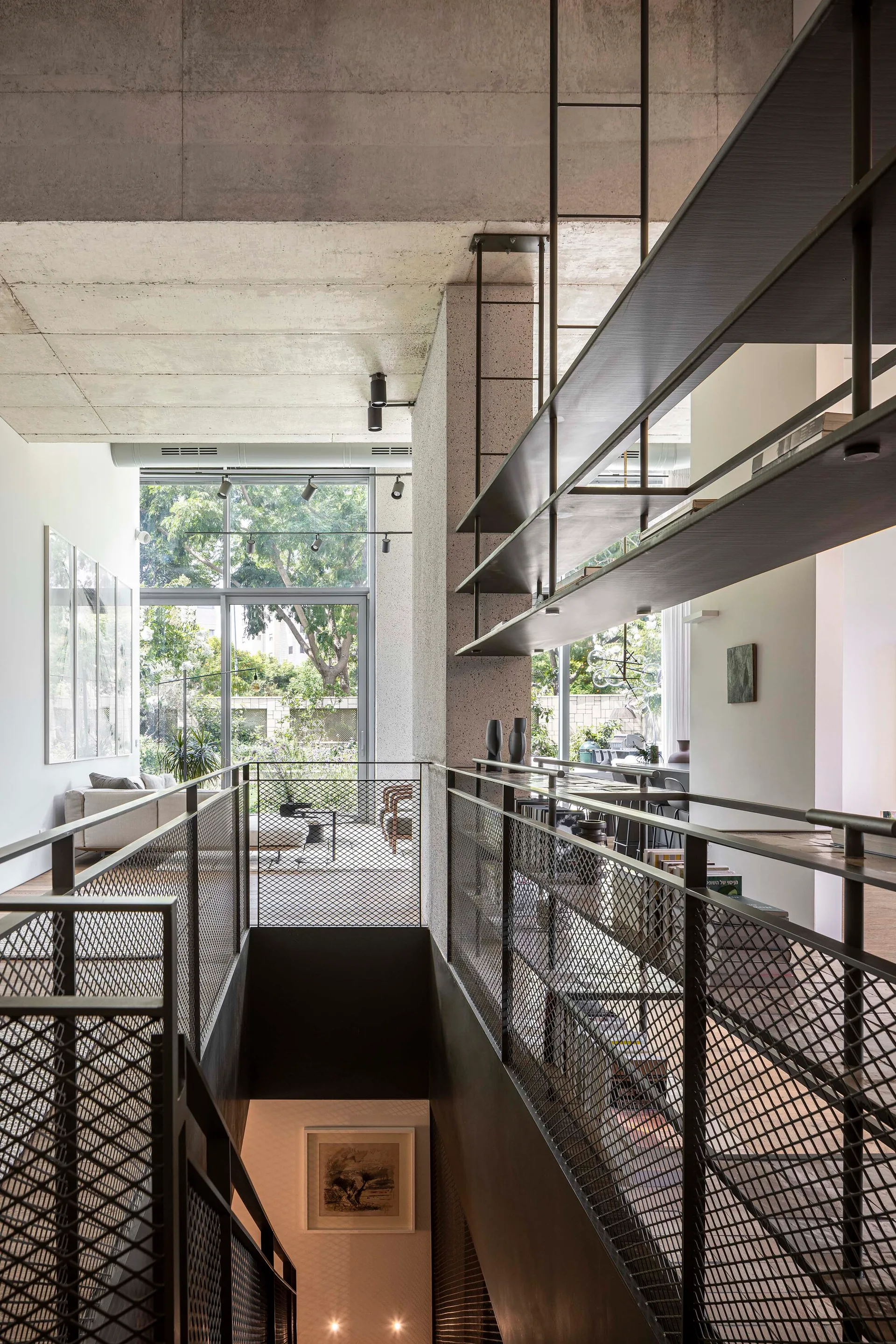 An industrial interior landing features black metal mesh railings, suspended shelving, and a raw concrete ceiling overlooking a lower level.