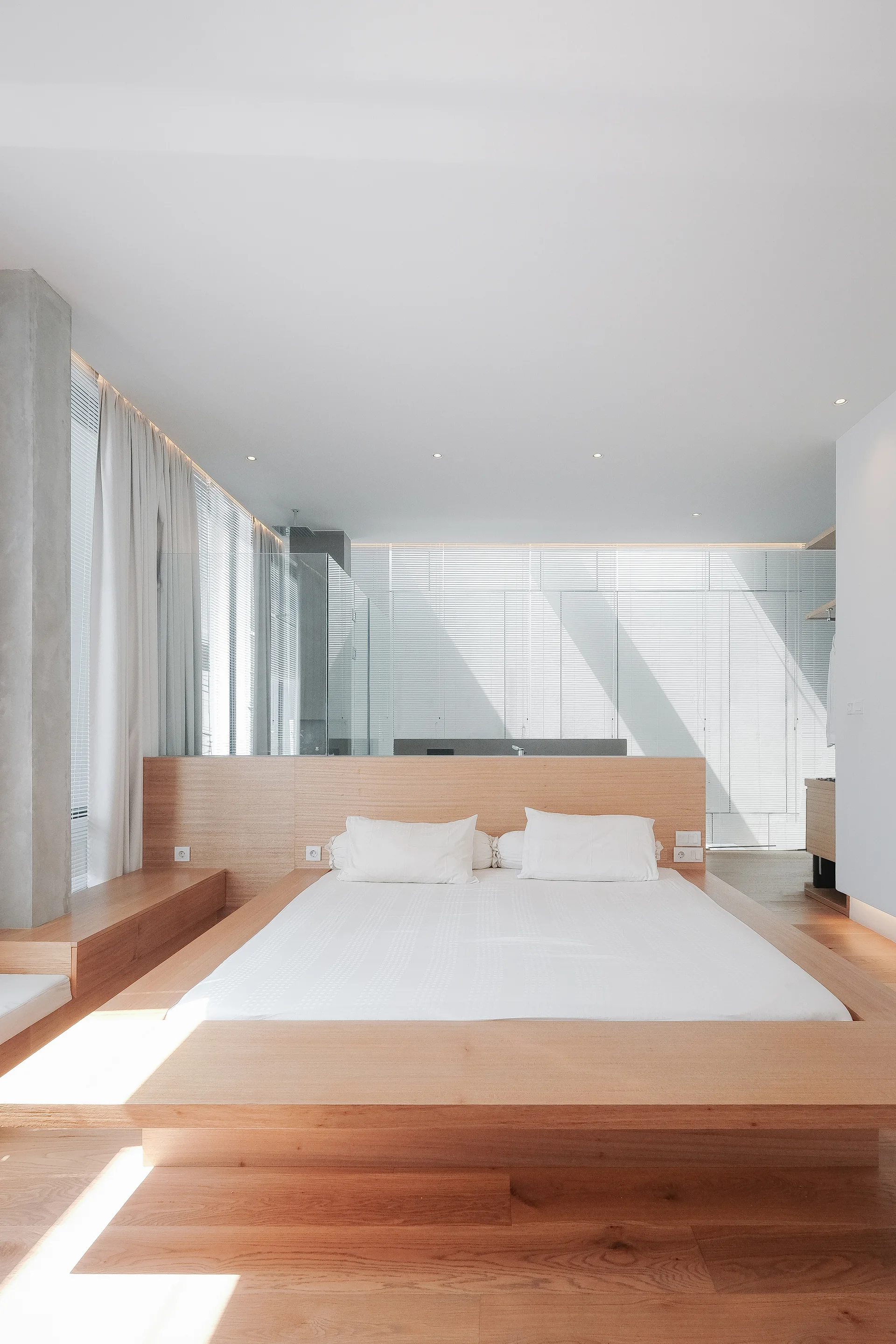 A low light wood platform bed sits against a matching wood headboard partition in a minimalist white bedroom.