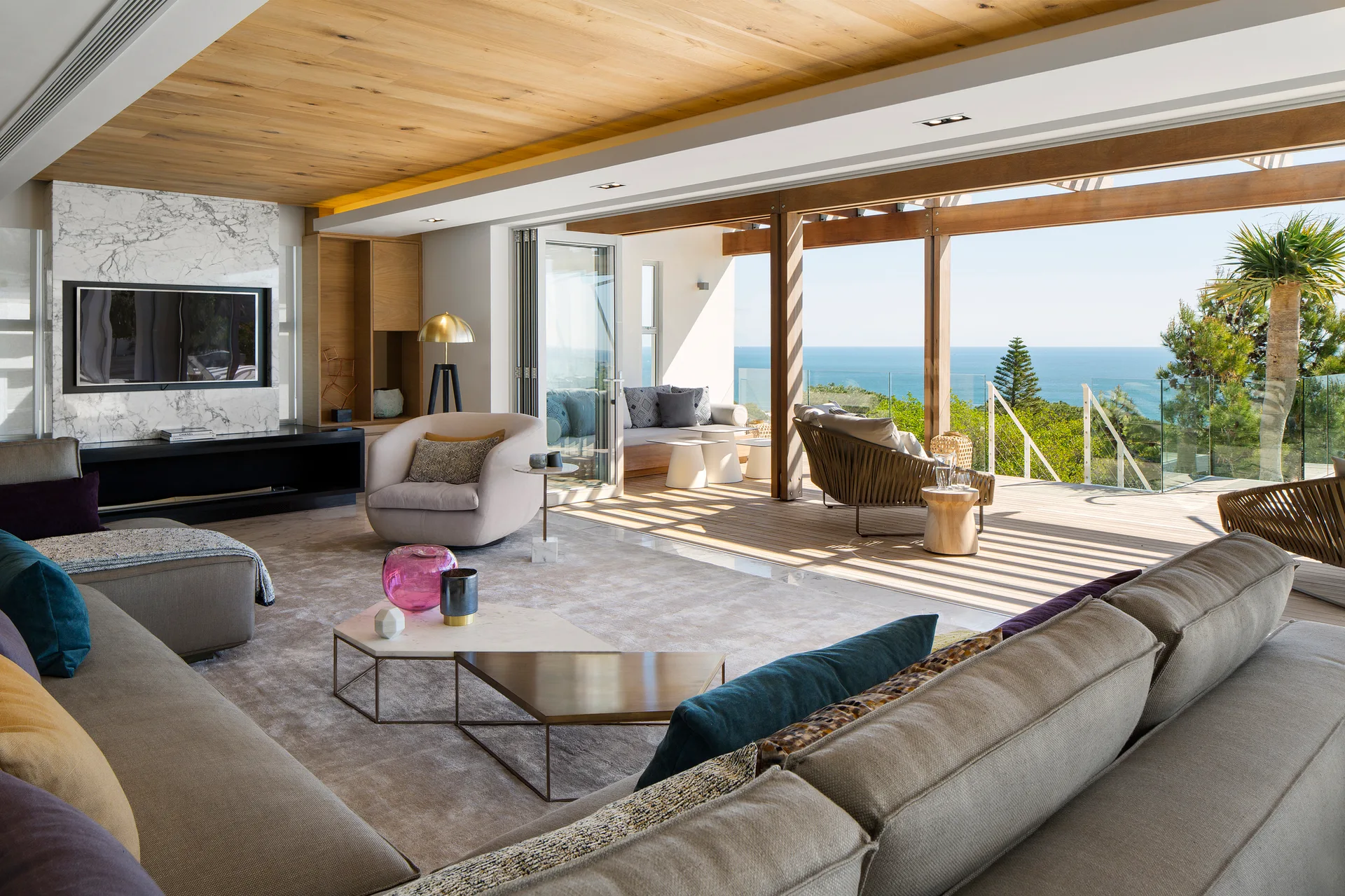 A modern living room features a tan sectional sofa, a marble media wall, and sliding doors opening to an outdoor deck with ocean views.