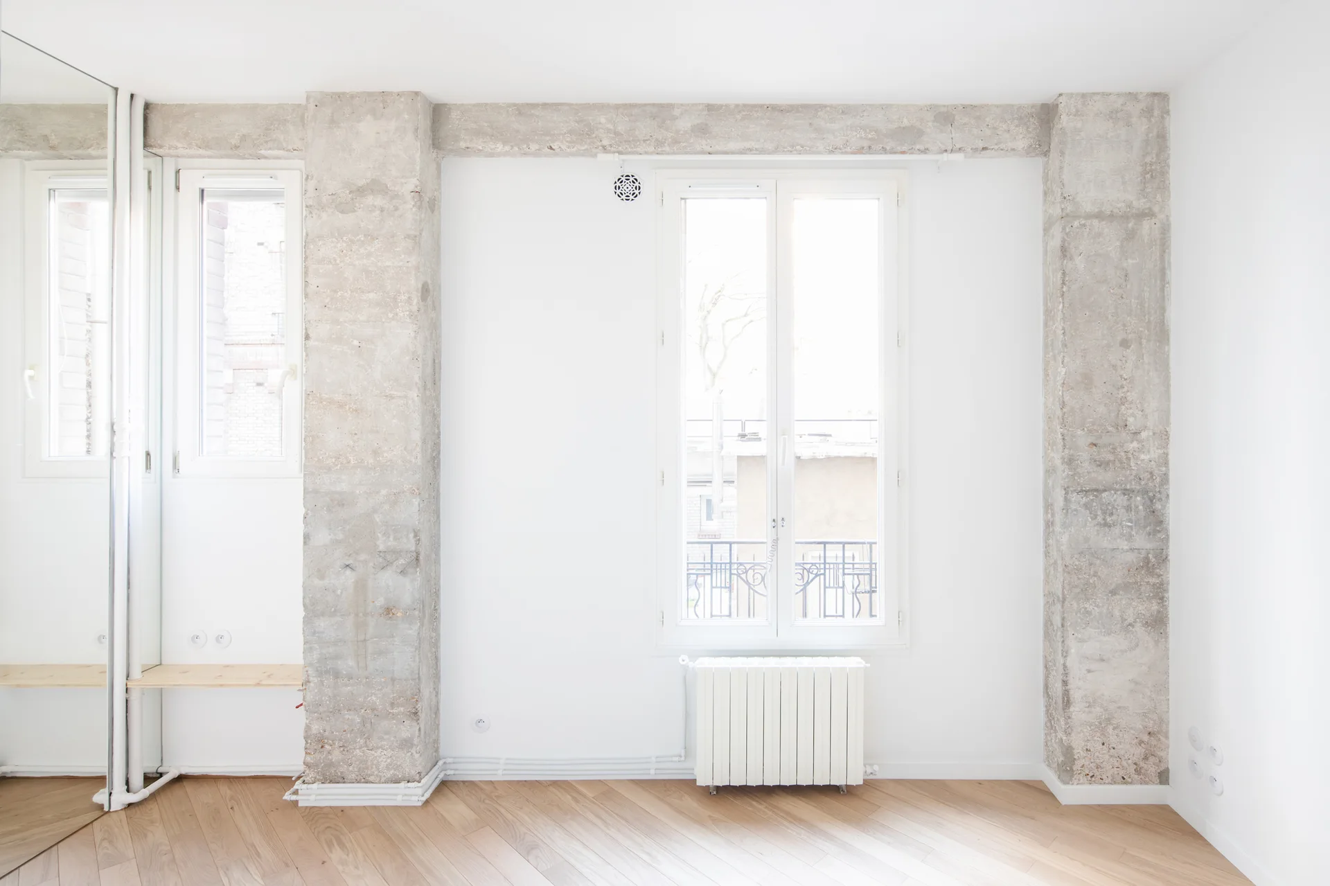 A bright minimalist room featuring raw concrete structural columns framing a tall white window and light-colored diagonal wood flooring.