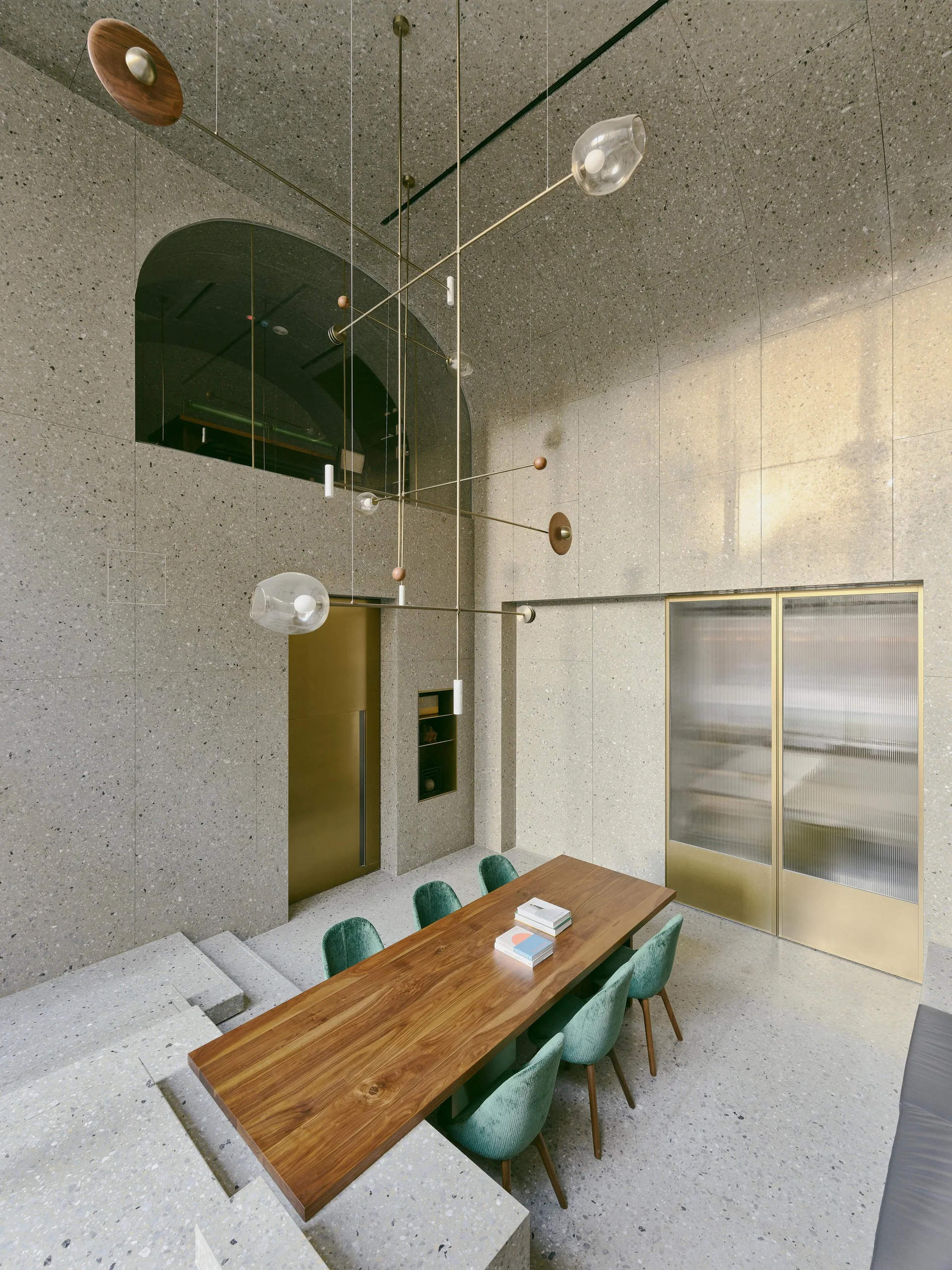 A spacious terrazzo-clad dining room featuring a long wooden table with green upholstered chairs beneath a brass chandelier and vaulted ceiling.