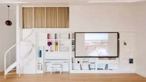 A white custom built-in shelving unit with a television and integrated desk sits next to a white metal spiral staircase on hardwood flooring.