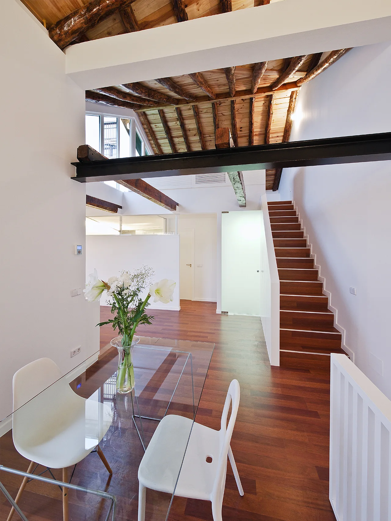 A modern open-plan dining area features a glass-topped table, white chairs, a wooden staircase, and exposed ceiling rafters with a steel beam.