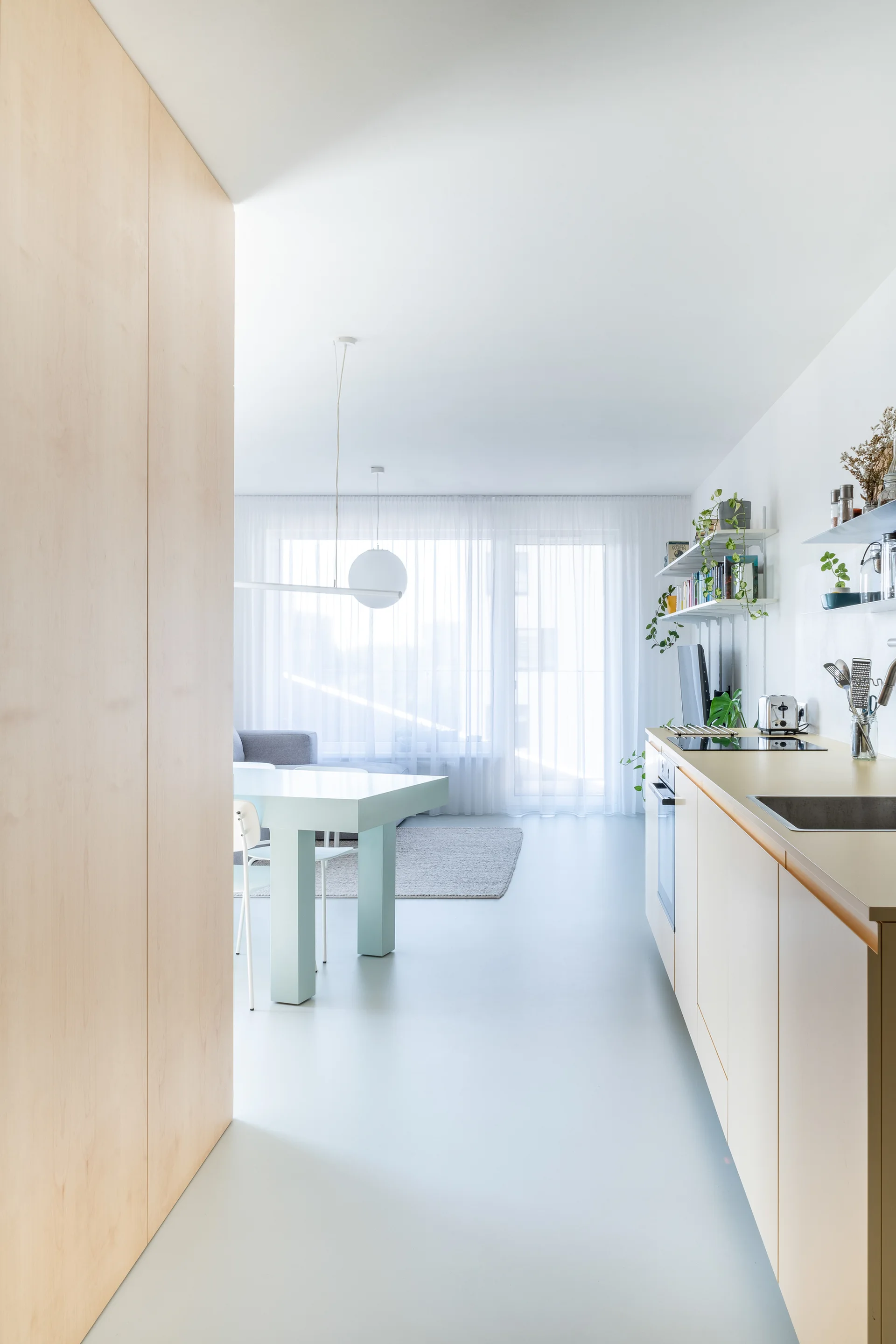 A minimalist open-plan kitchen and dining area featuring flat-panel light wood cabinets, a pale mint green dining table, and sheer white curtains.