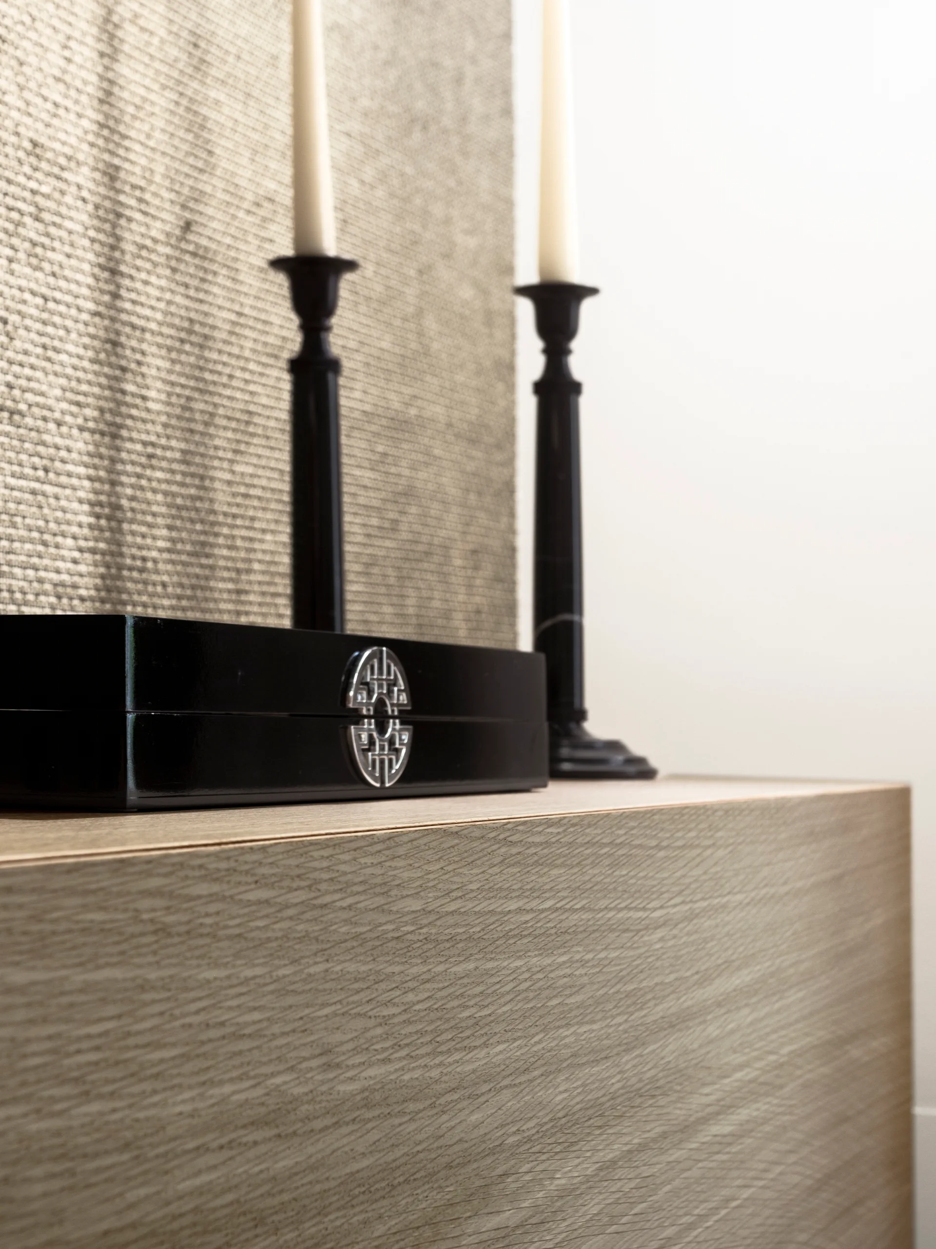 A black lacquered decorative box and two tall black candlesticks rest on a light wood console table against a textured woven background.