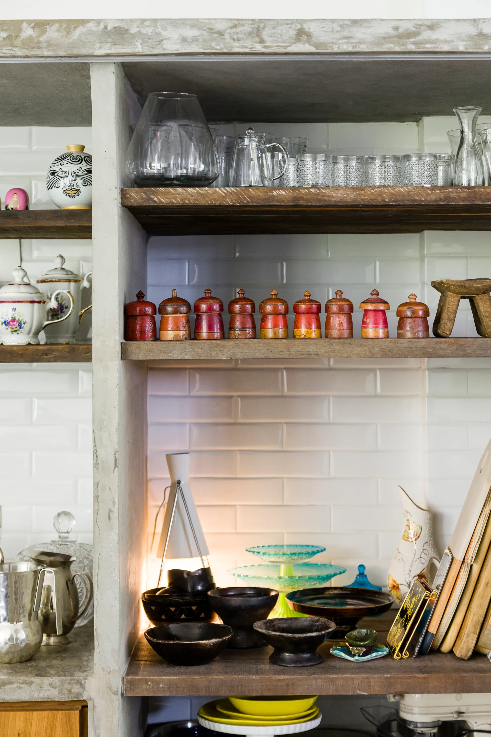 Rustic wooden shelves framed by concrete against a white subway tile backsplash feature glassware, colorful small containers, and a table lamp.