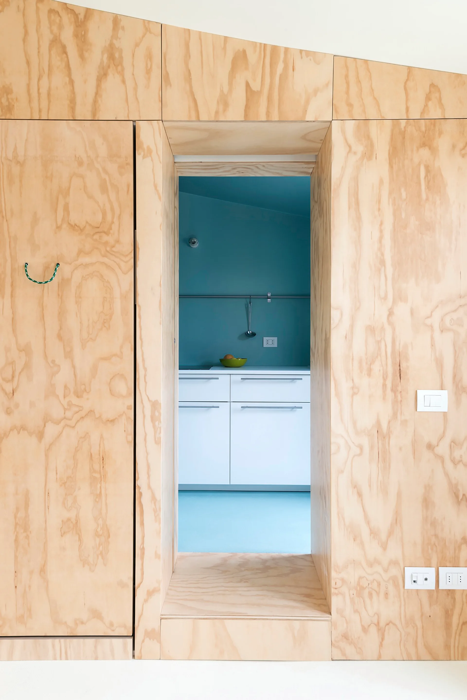 A view through a deep doorway framed in light plywood paneling, revealing a kitchen with white cabinets and teal blue walls.