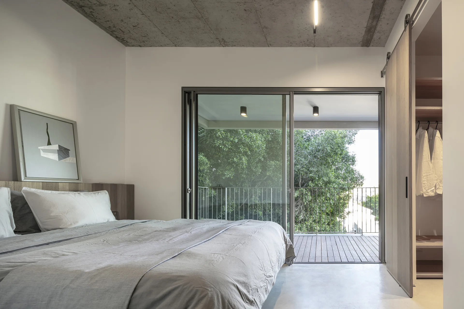 Minimalist bedroom featuring an exposed concrete ceiling, a grey bed, a sliding glass door to a balcony, and a wood-toned sliding closet door.