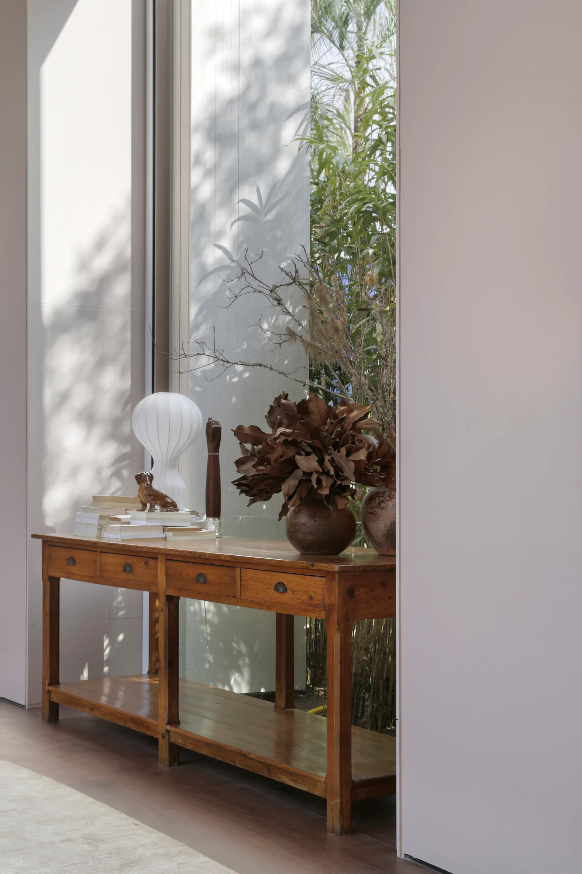 A long wooden console table with drawers stands in front of a tall window, holding a white table lamp, stacked books, and vases of dried foliage.