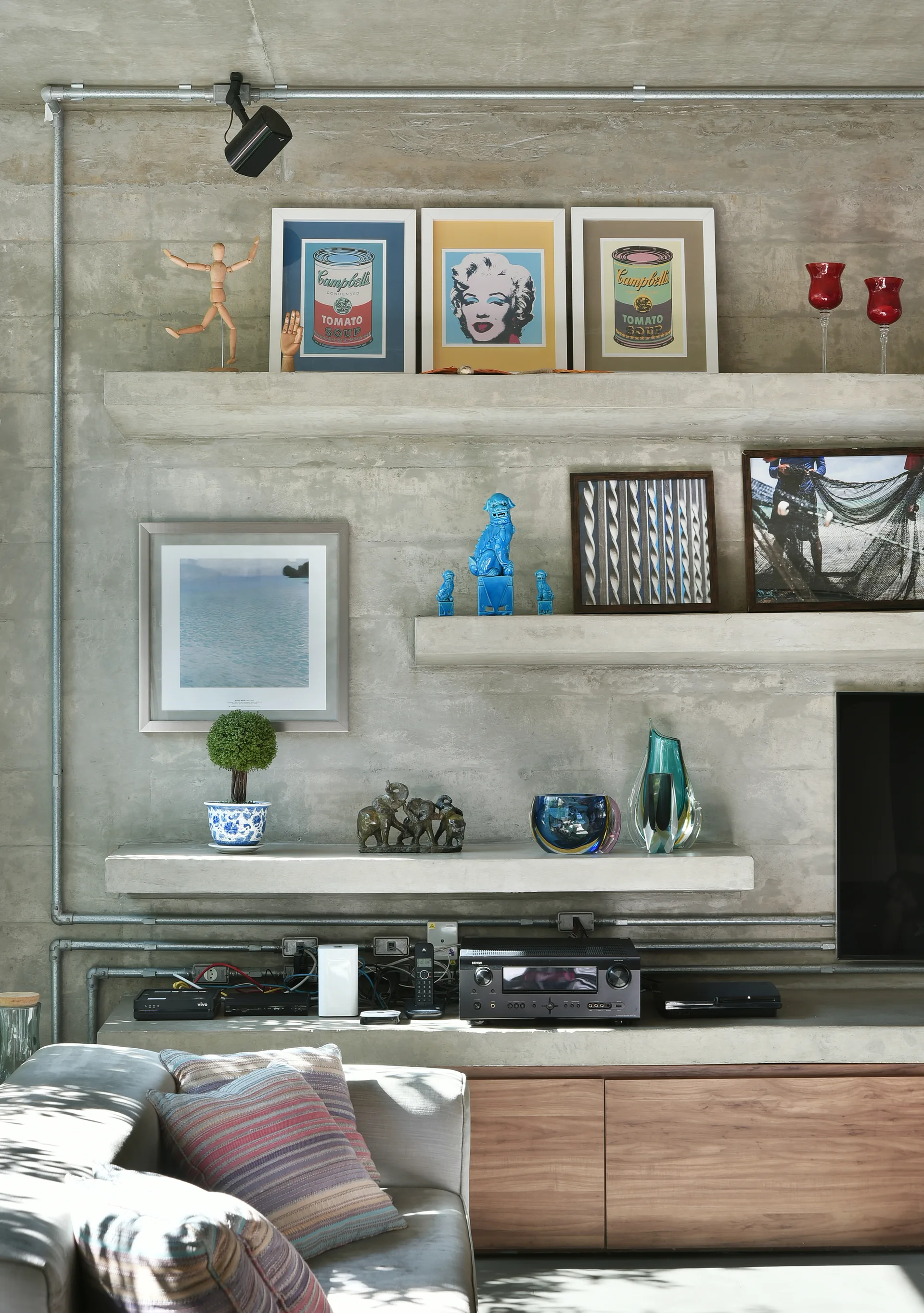 Concrete floating shelves on a raw concrete wall display colorful pop art and sculptures above a media console and grey sofa.
