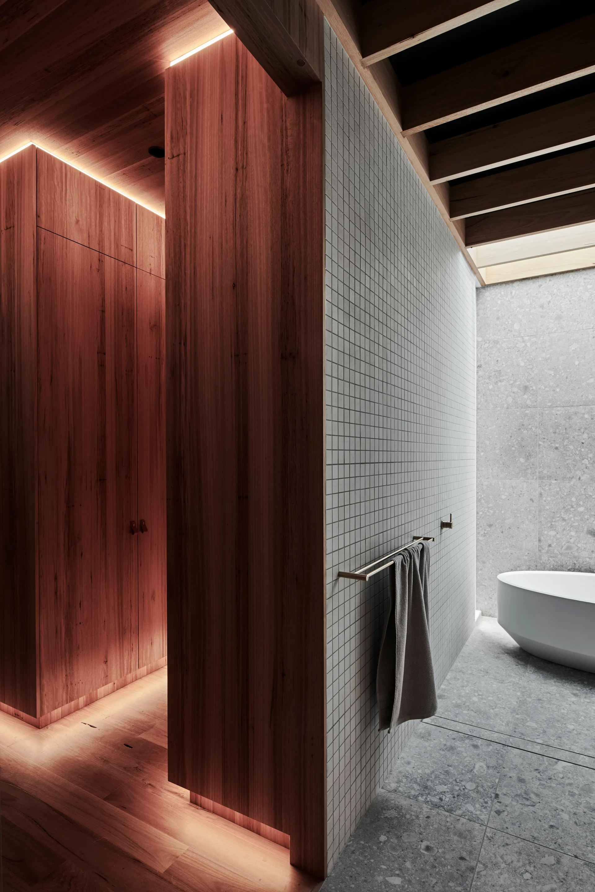 A modern minimalist bathroom featuring warm wood paneling with integrated lighting, a white grid-tiled wall, and a freestanding white bathtub.