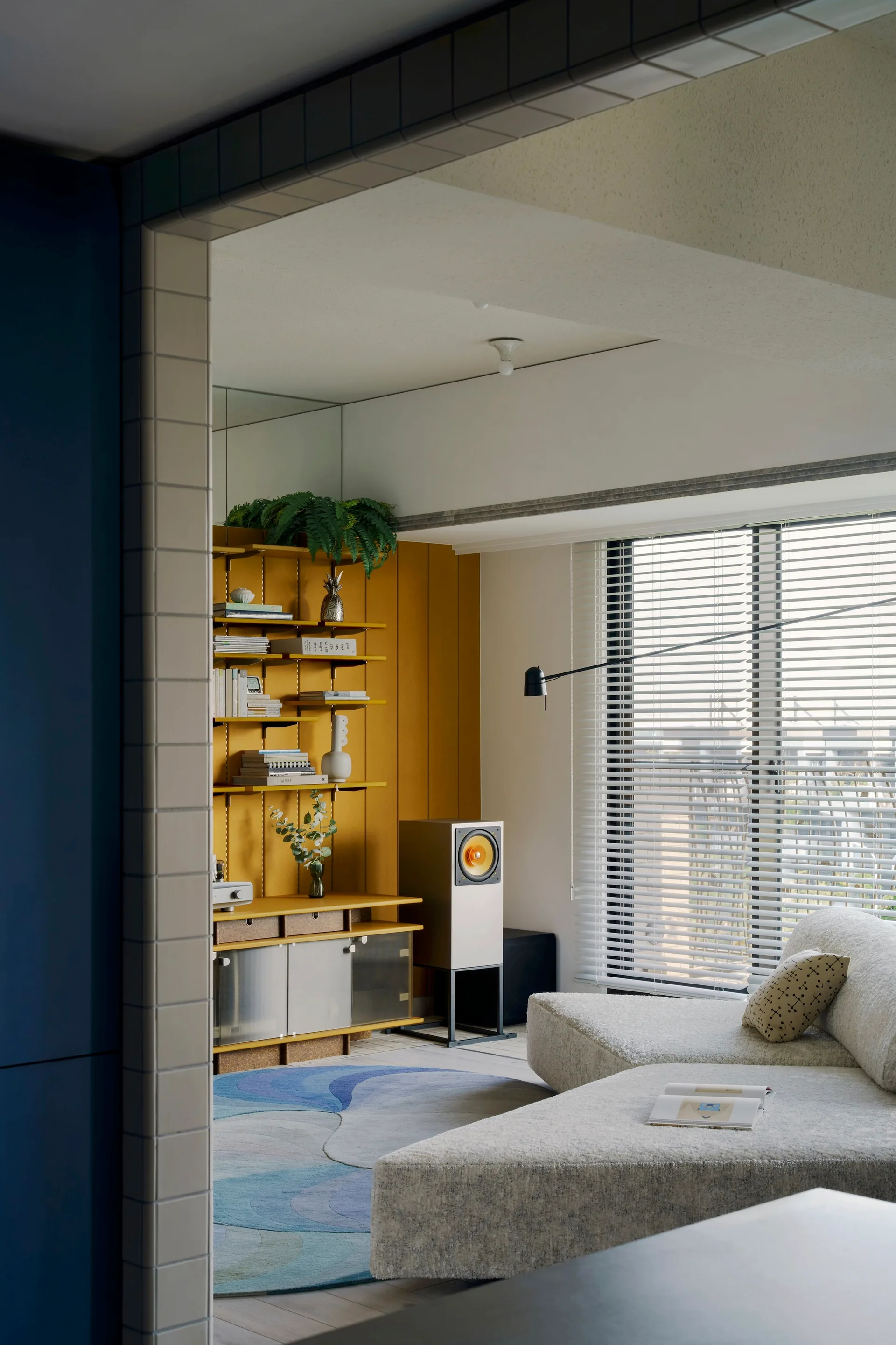A living room seen through a tiled doorway features a textured white modular sofa, a blue patterned rug, and a mustard yellow wall shelf.