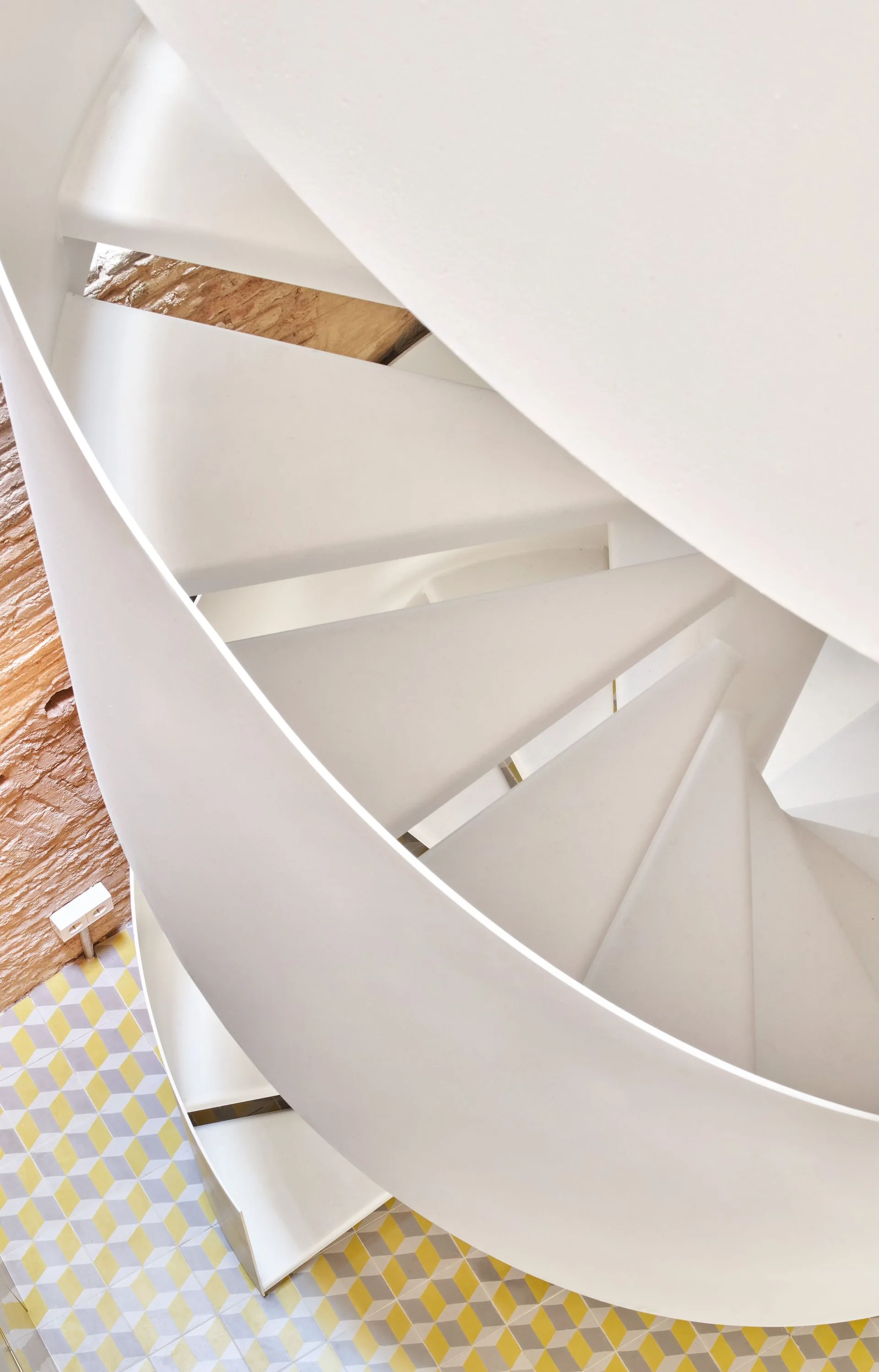 An overhead view of a curved white metal spiral staircase leading down to yellow and grey geometric tiled flooring.