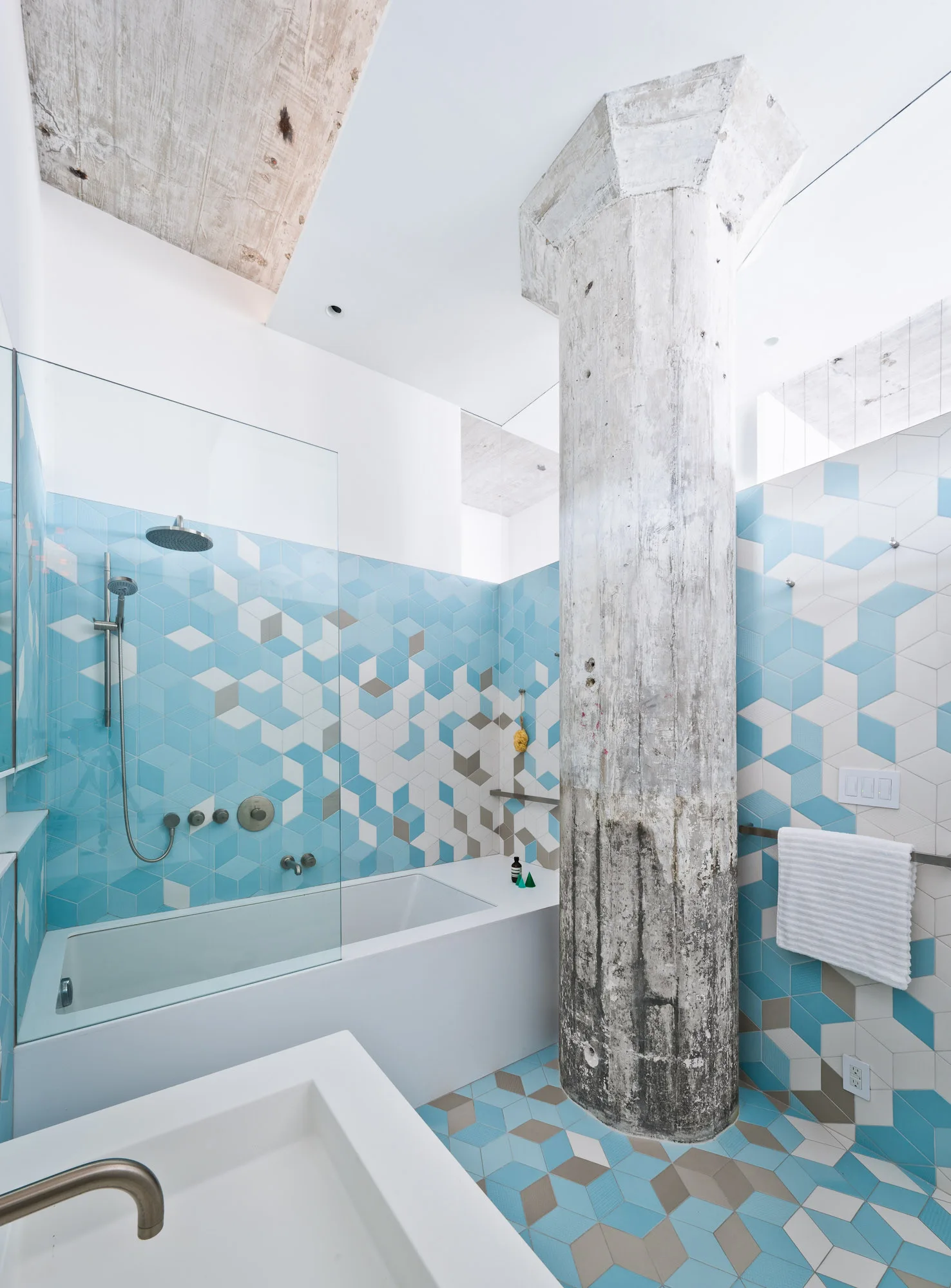 A bathroom features blue, grey, and white hexagonal geometric tile walls and floors, a white bathtub, and a large weathered concrete column.