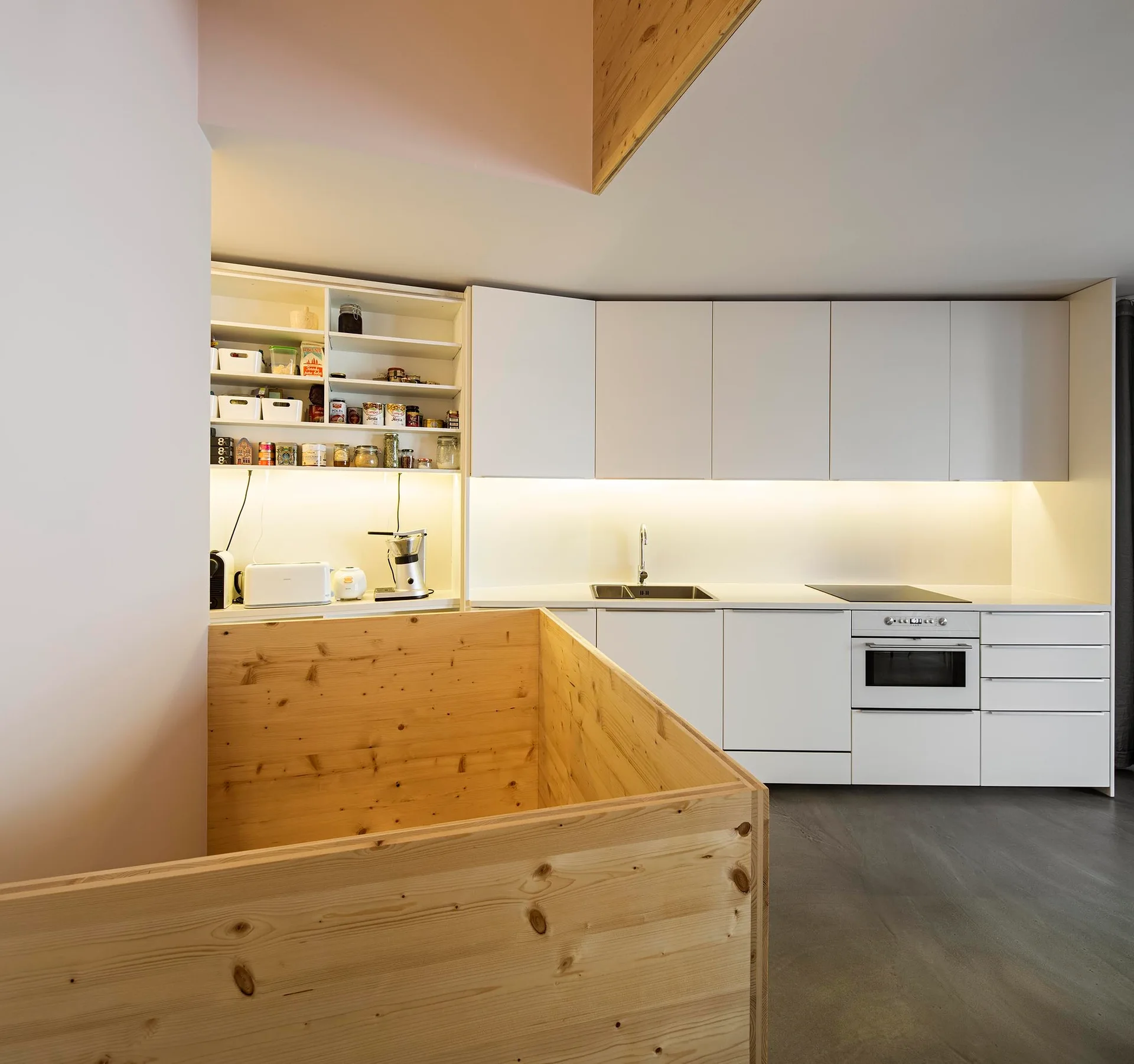 A minimalist kitchen features flat-front white cabinets, integrated under-cabinet lighting, and a light pine wood staircase enclosure.