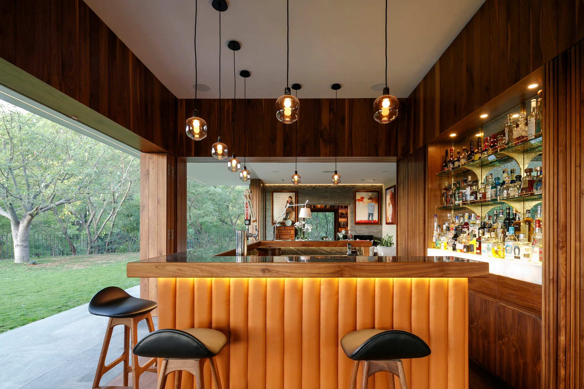 An indoor home bar features a wood counter with an orange fluted front, black stools, and glass pendant lights looking out to a green garden.