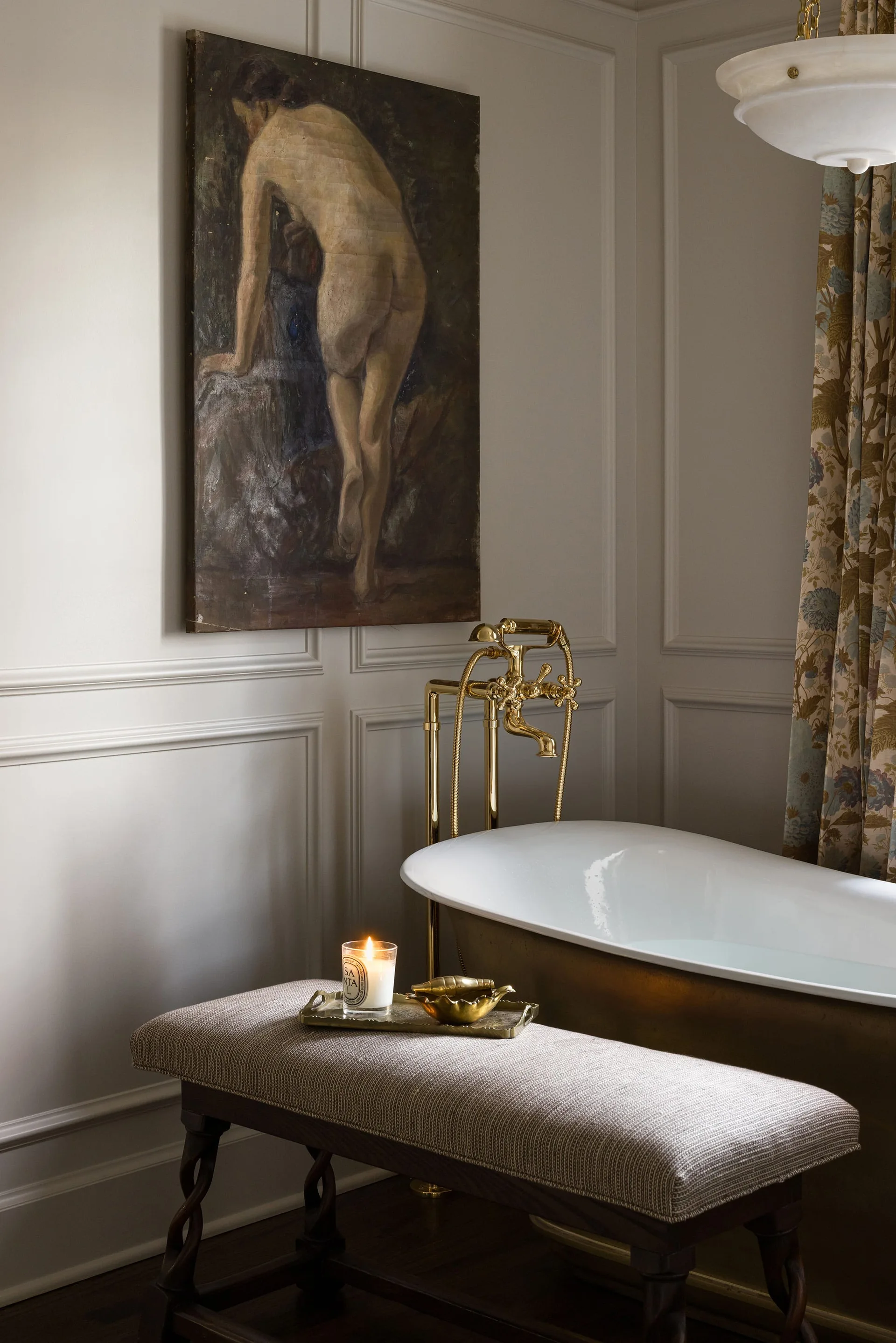 A classic bathroom featuring a bronze freestanding bathtub, gold floor-mounted faucet, gray paneled walls, and an upholstered bench with a candle.