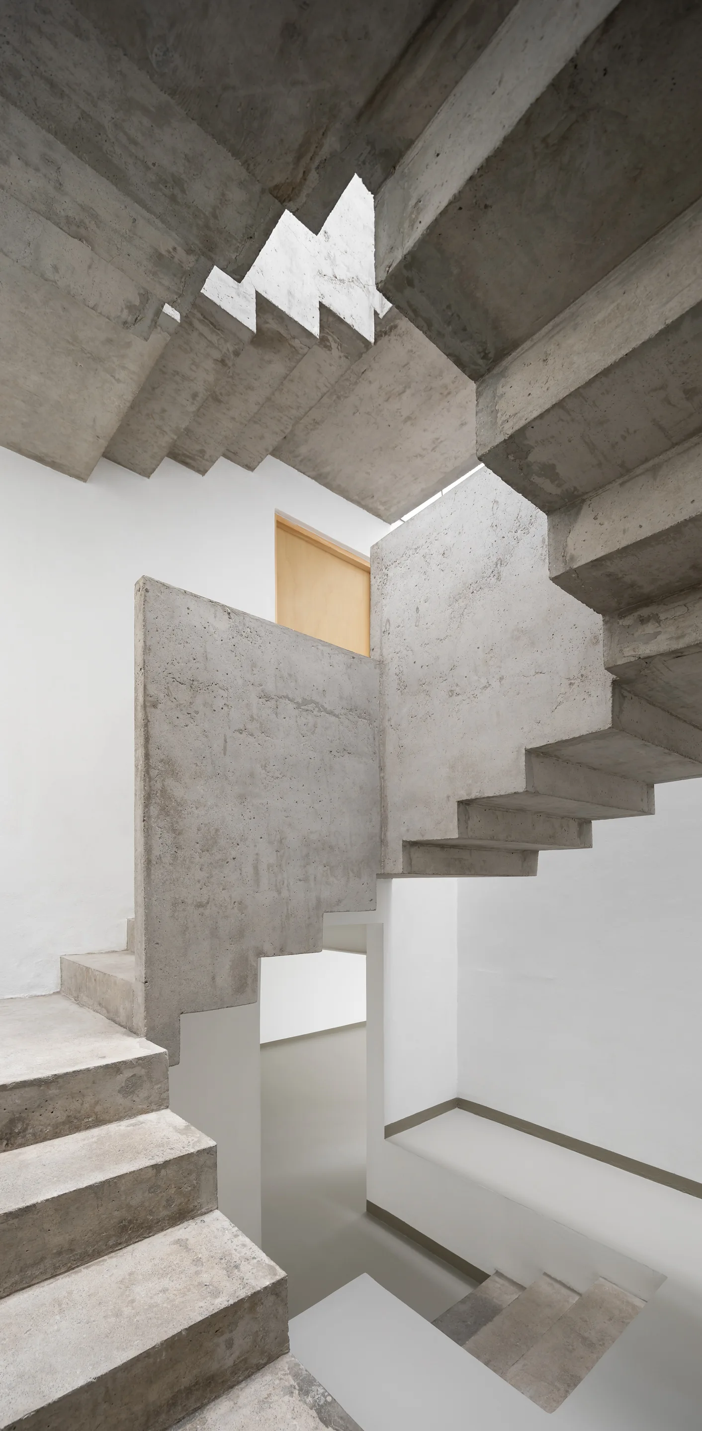 A multi-level staircase constructed of raw grey concrete winding through a minimalist interior with smooth white walls and a single wood door frame.