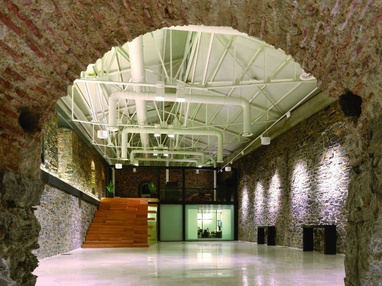 An expansive industrial hall viewed through a historic stone archway features exposed stone walls, a white truss ceiling, and a wood staircase.
