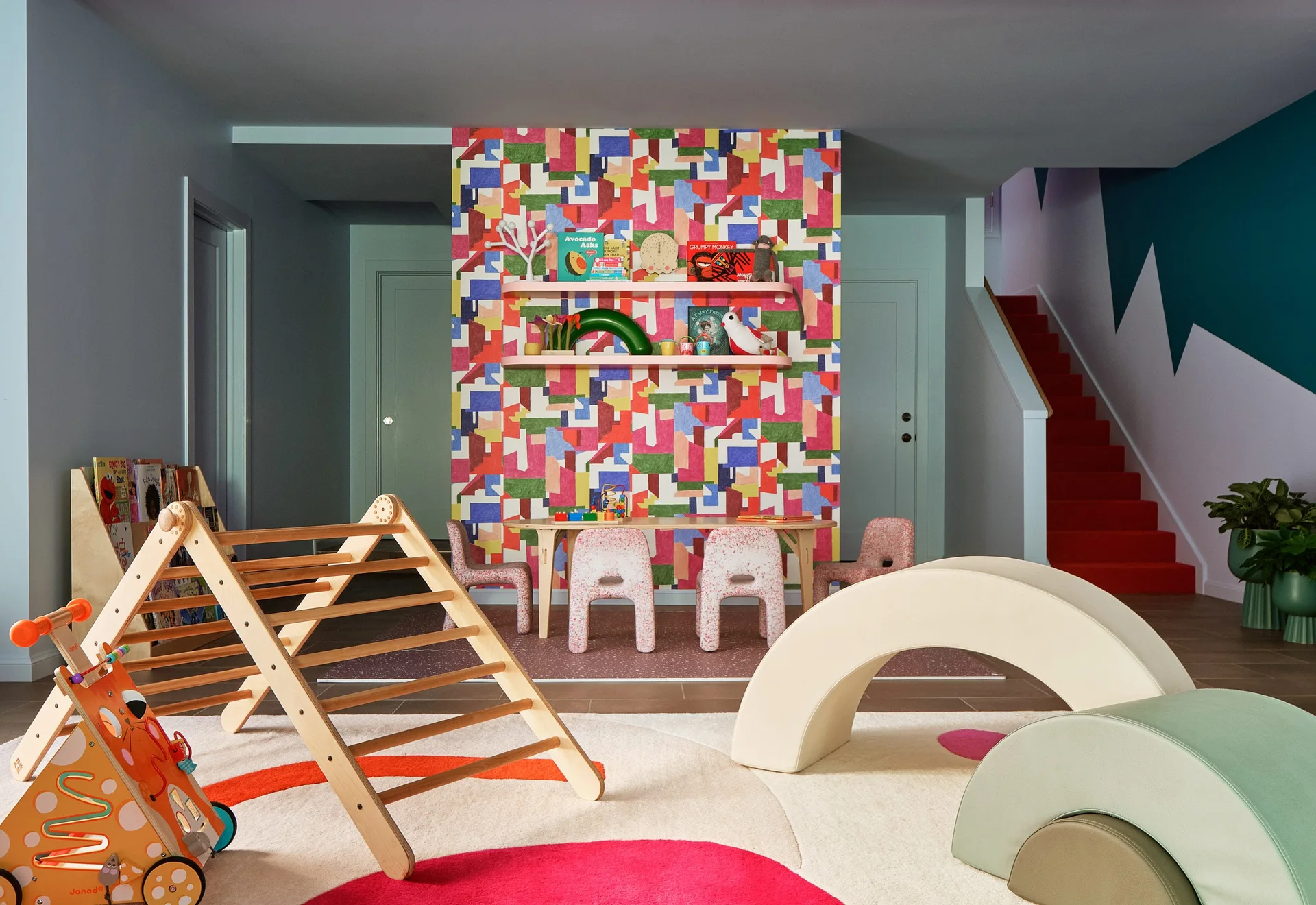 A children's playroom features a colorful geometric accent wall, a wooden climbing triangle, foam play shapes, and a small table set.