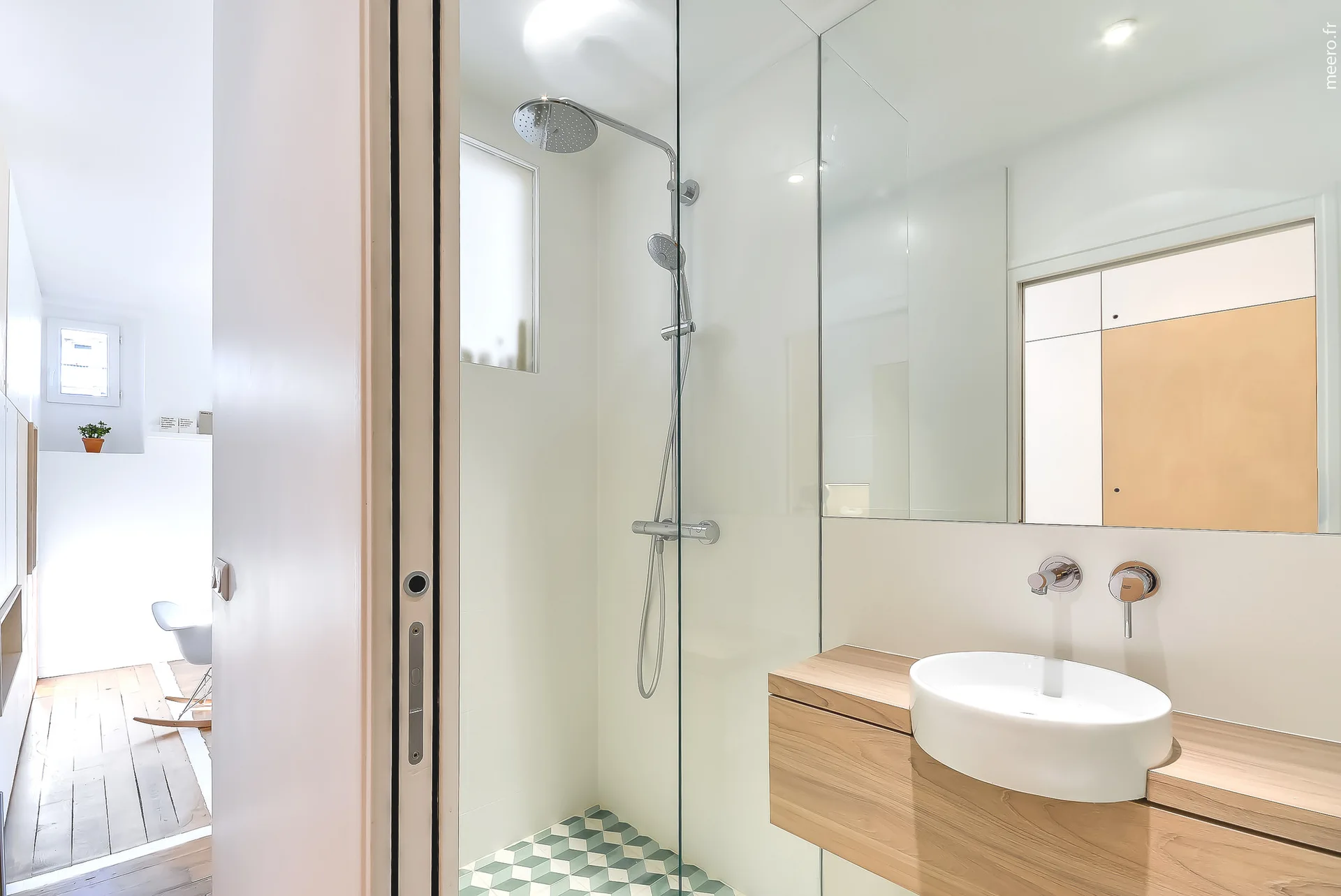 A minimalist bathroom with a floating wood vanity, circular white vessel sink, and a glass shower stall featuring green geometric floor tiles.