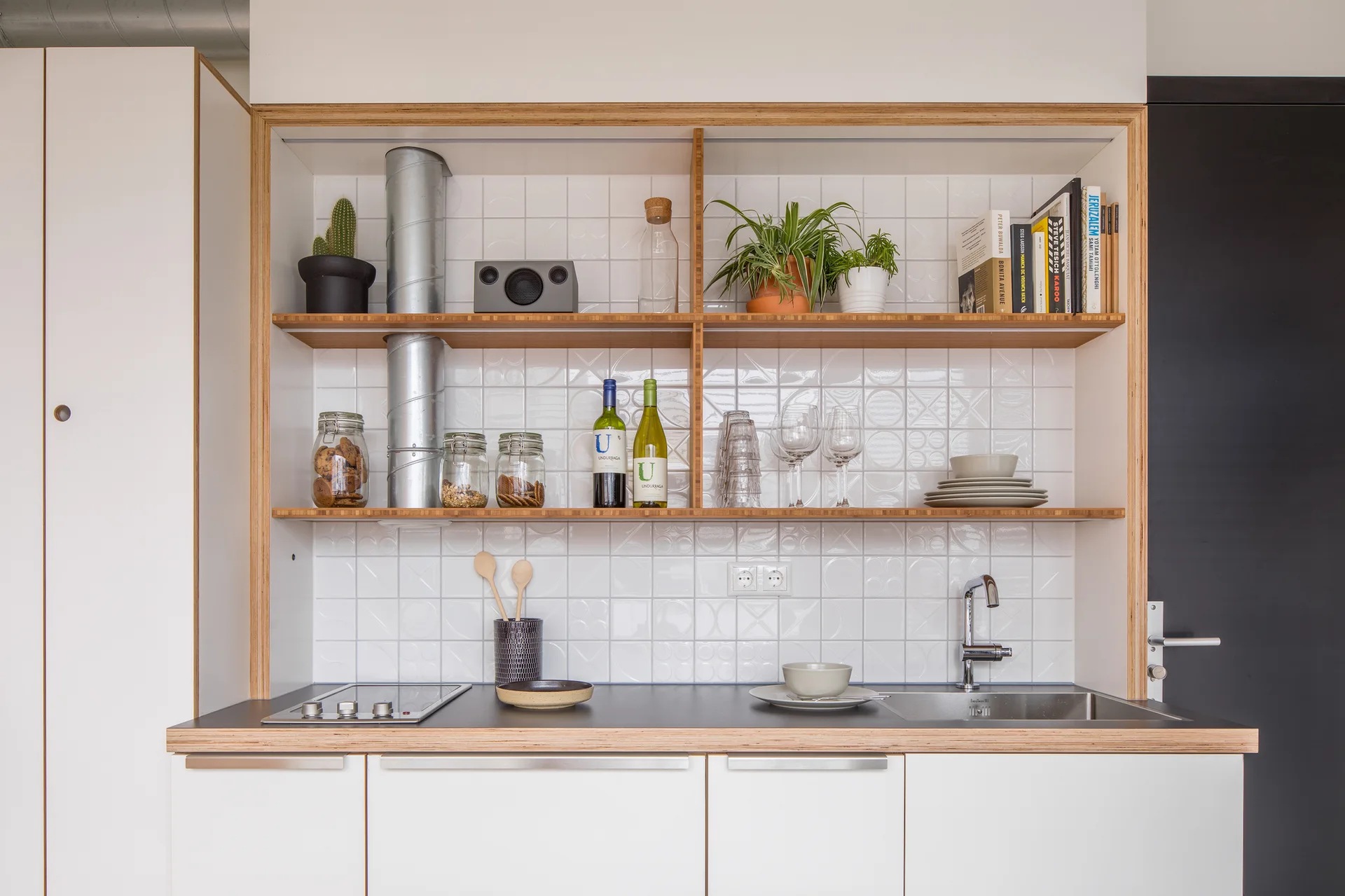 A compact kitchenette features white base cabinets, a black countertop with a sink, and wooden open shelving against a white geometric tile backsplash.