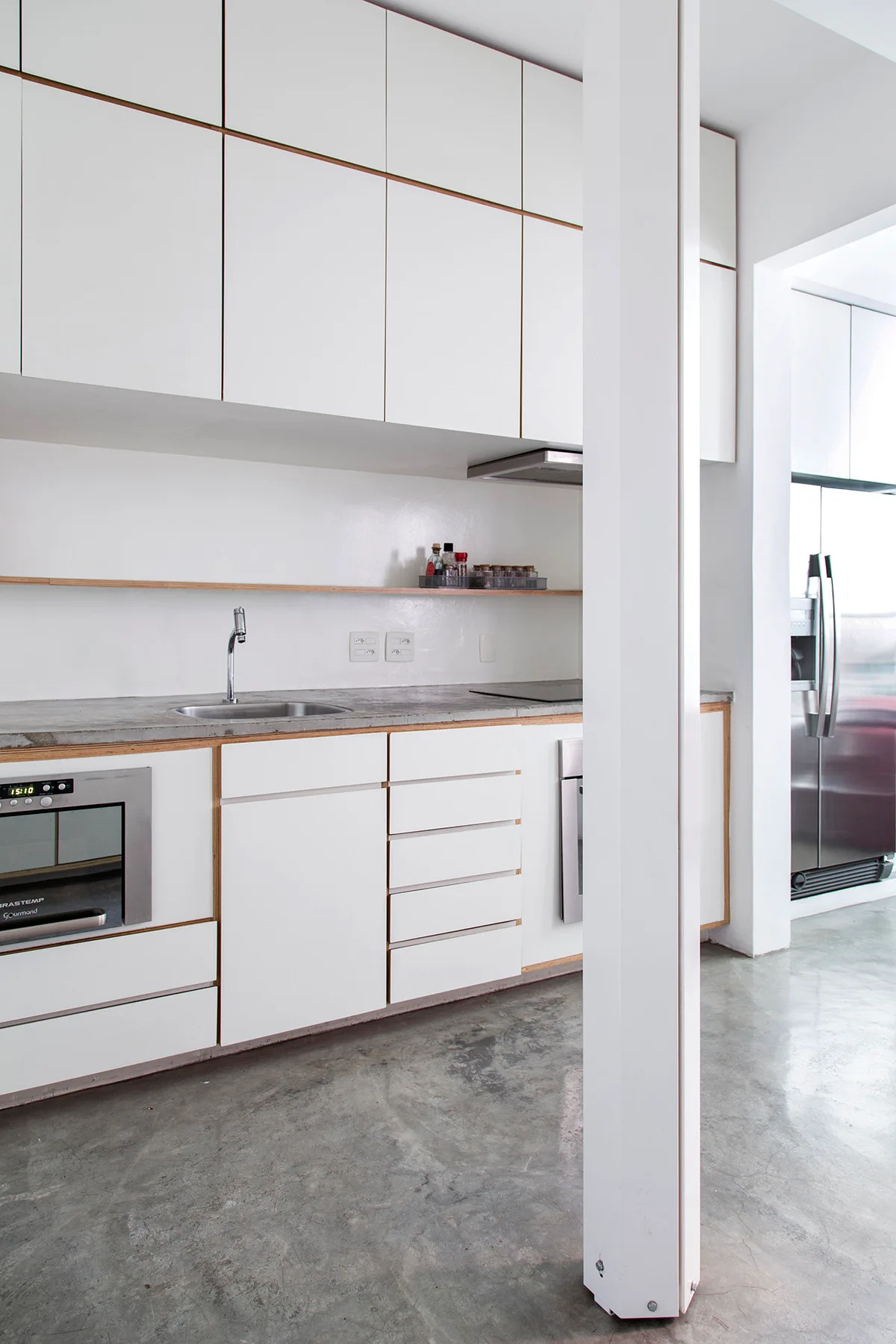 A minimalist white kitchen features flat-front cabinets with exposed plywood edges, a grey stone countertop, and a polished concrete floor.