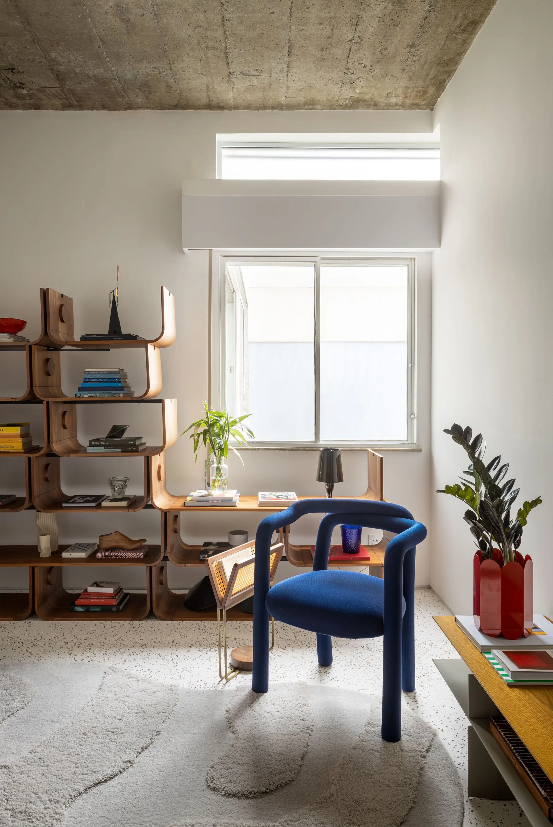 A modern living space featuring a cobalt blue accent chair, a curved wood modular shelf, speckled terrazzo flooring, and an exposed concrete ceiling.