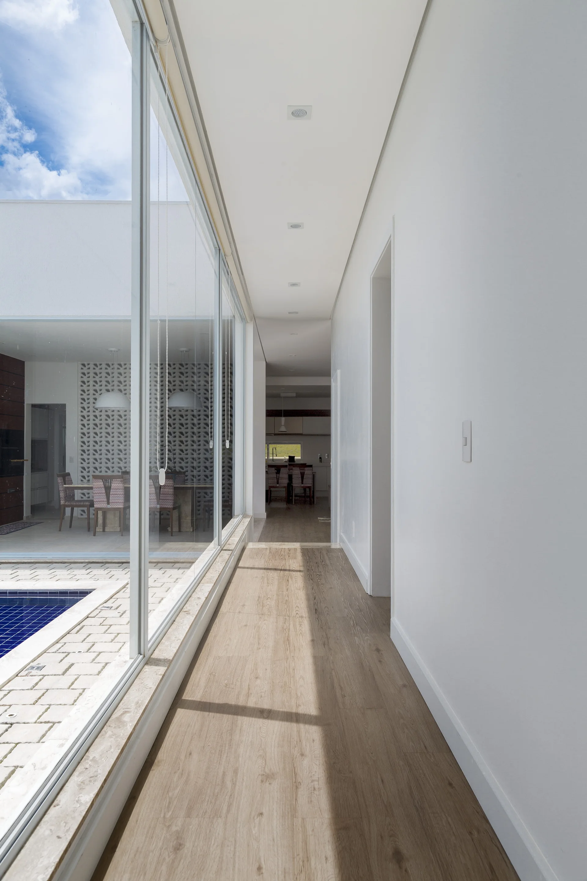 A long white residential hallway with light wood flooring features a floor-to-ceiling glass window wall overlooking an outdoor swimming pool.