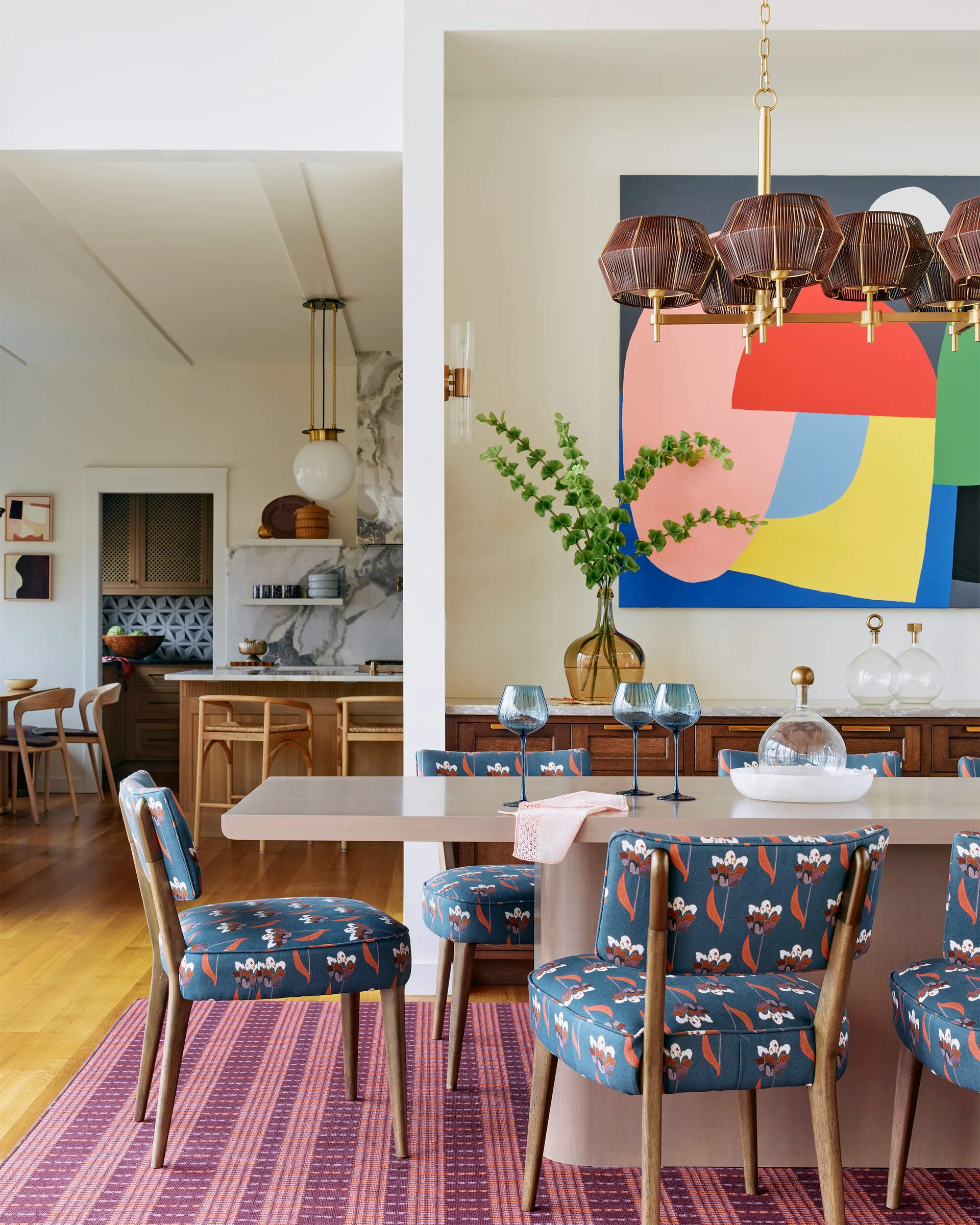 A dining room features blue floral patterned chairs around a light wood table, set on a textured purple rug beneath a large abstract painting.