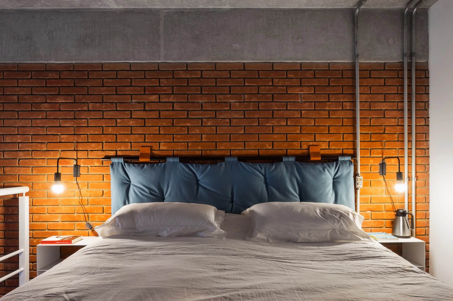 A bedroom with an exposed brick wall, a hanging blue headboard on the bed, and black wall sconces under a concrete ceiling.