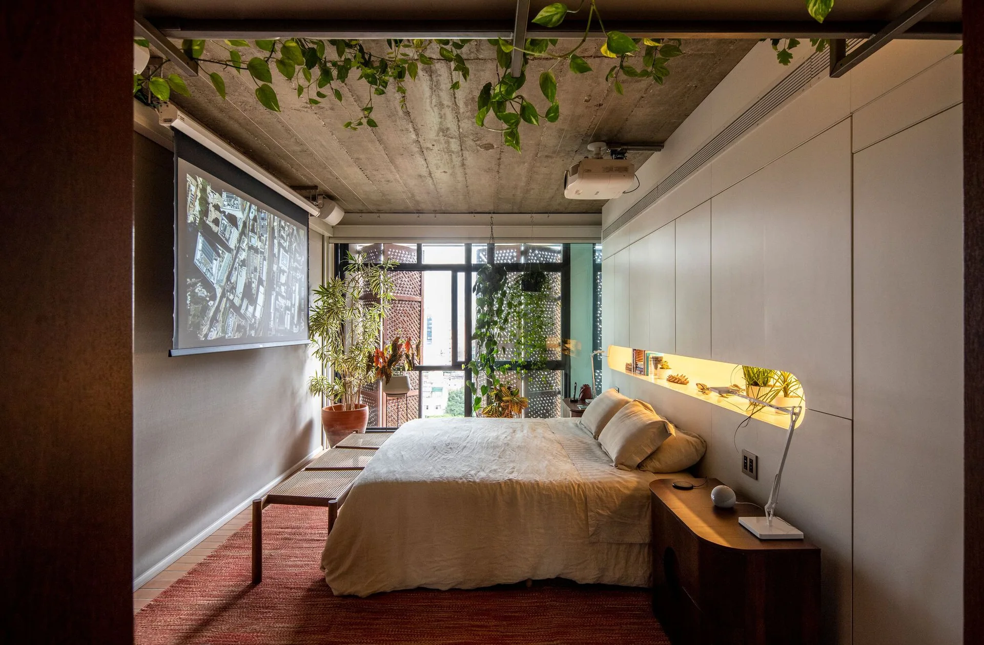 A minimalist bedroom features a central bed under an exposed concrete ceiling, flanked by a white built-in wall unit and a wall-mounted projector screen.