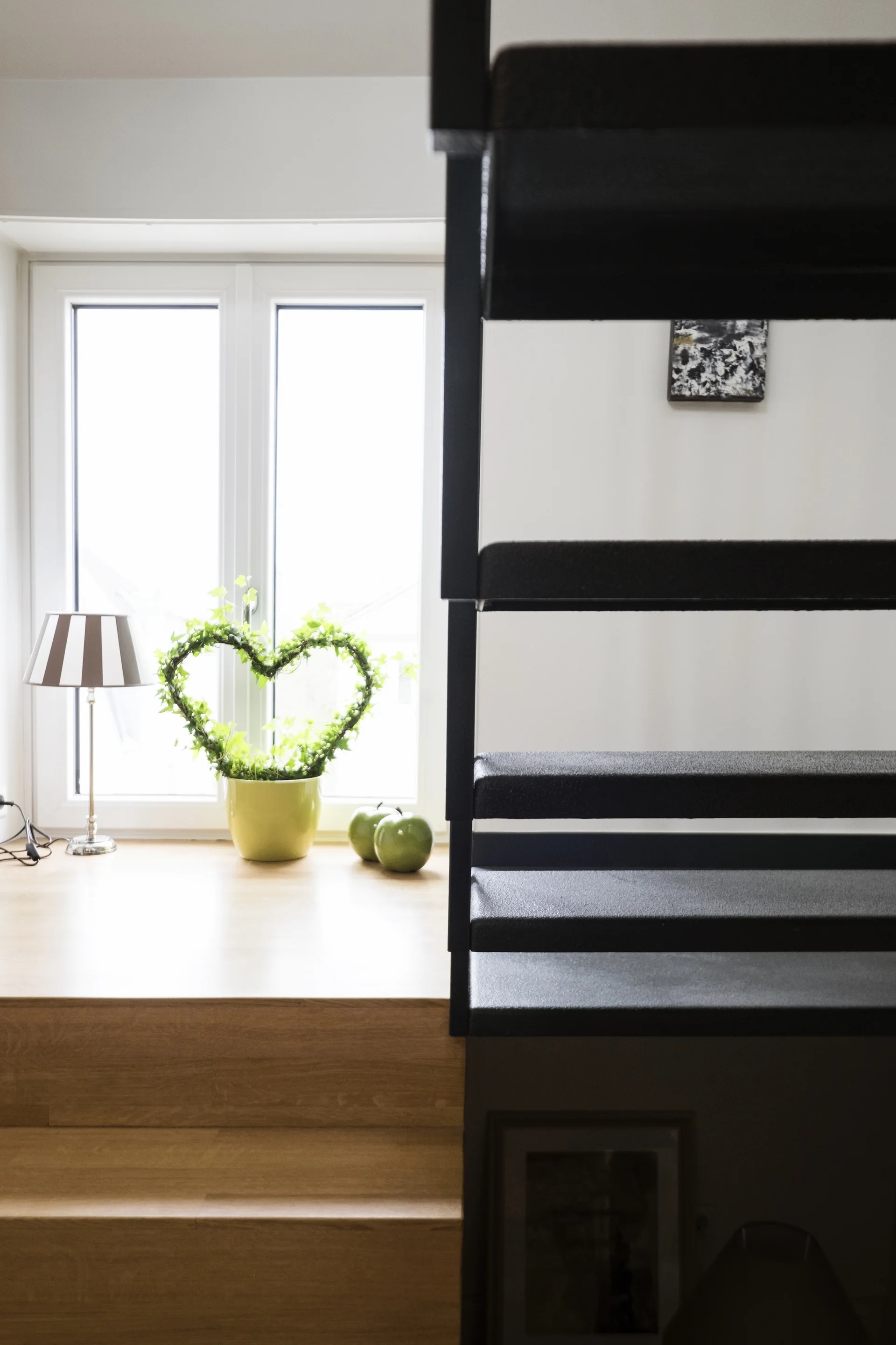 A black metal staircase stands next to a wooden landing with a bright window displaying a heart-shaped green plant and striped lamp.