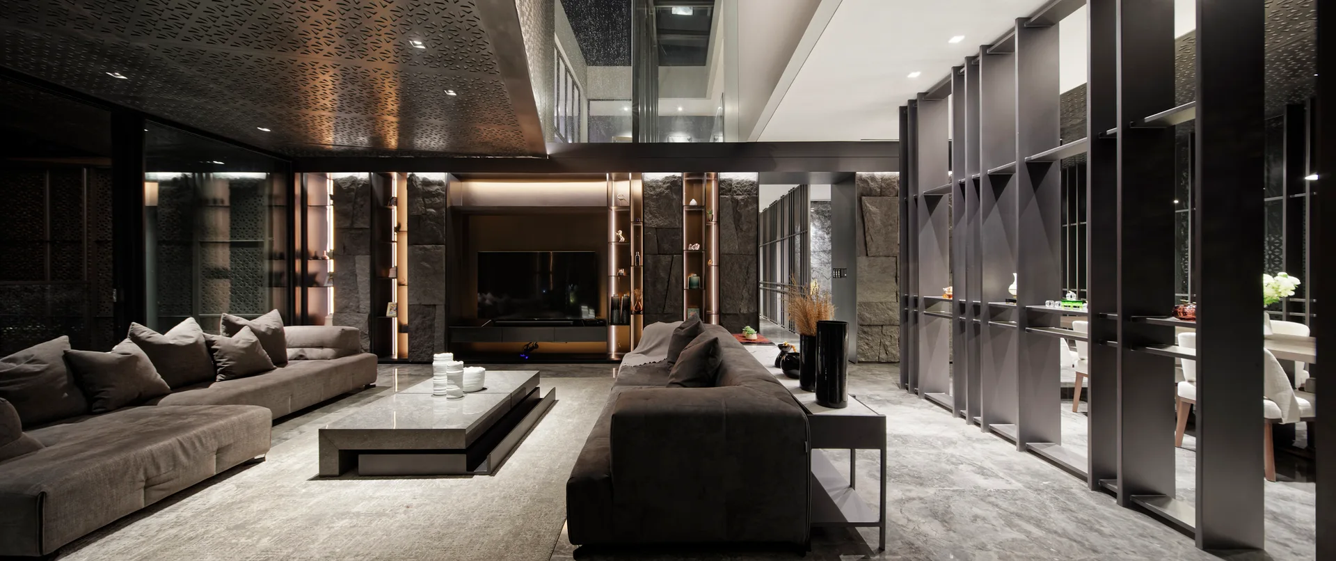 A spacious grey living room with a low-profile sectional sofa, stone coffee table, textured dark stone accent wall, and tall metal grid shelving.