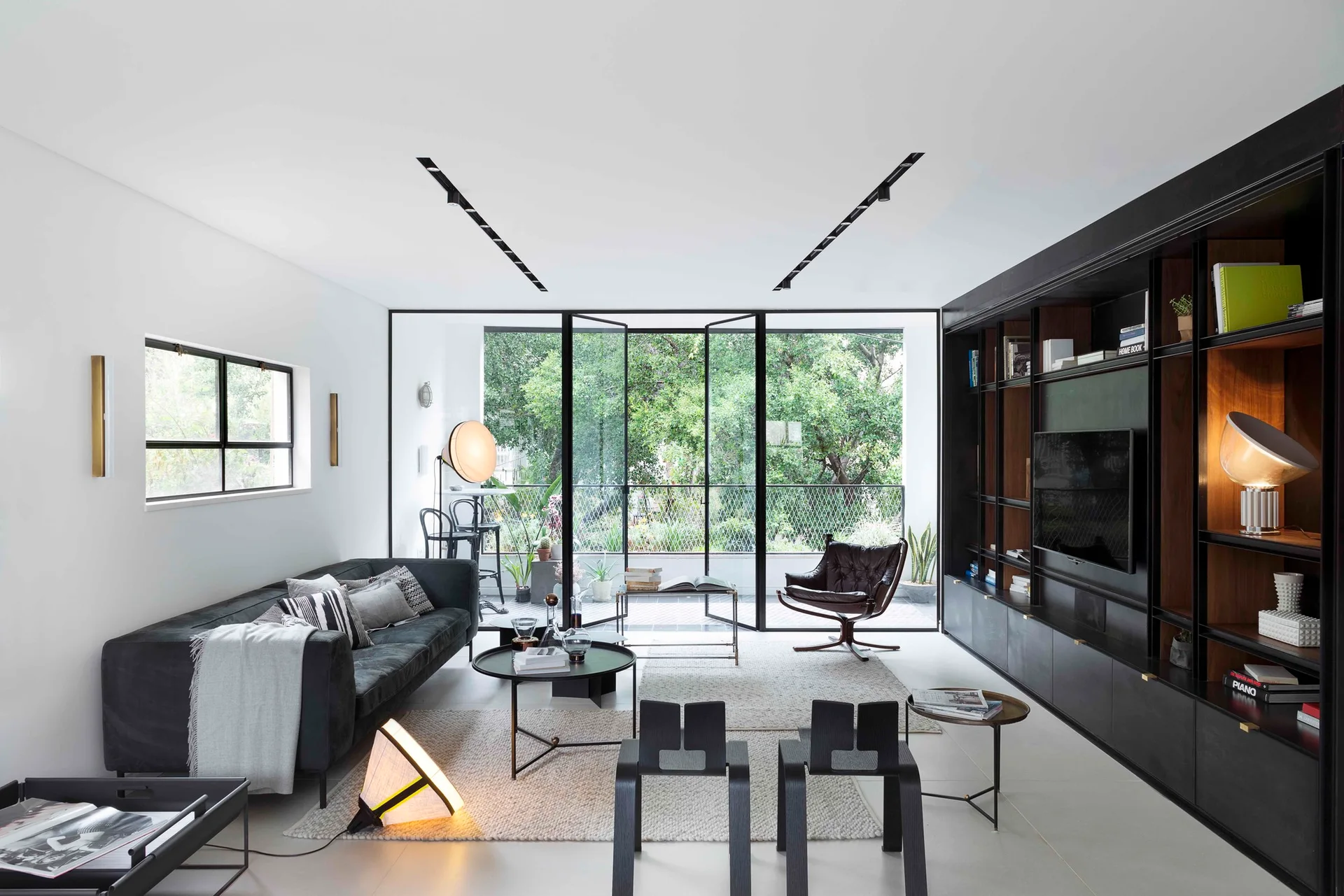 A modern living room features a large black shelving unit, a dark sofa, and steel-framed glass doors that open to a lush garden.