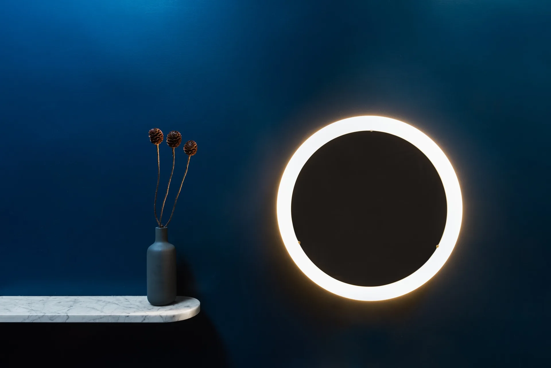 A glowing circular halo wall light is mounted on a deep navy blue wall next to a marble floating shelf with a small dark vase.