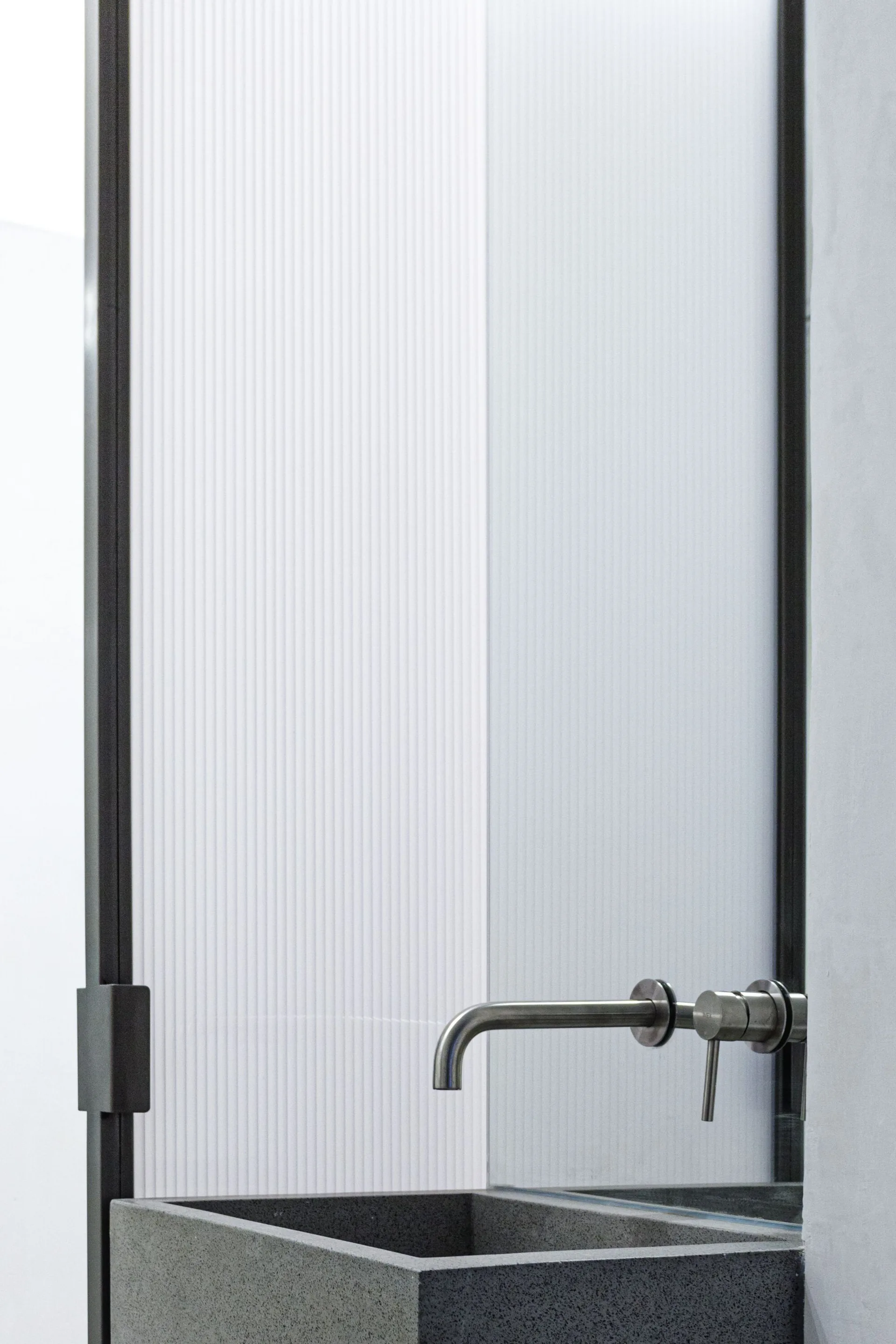 A wall-mounted metal faucet sits over a grey stone sink basin in front of a fluted frosted glass partition with a black metal frame.
