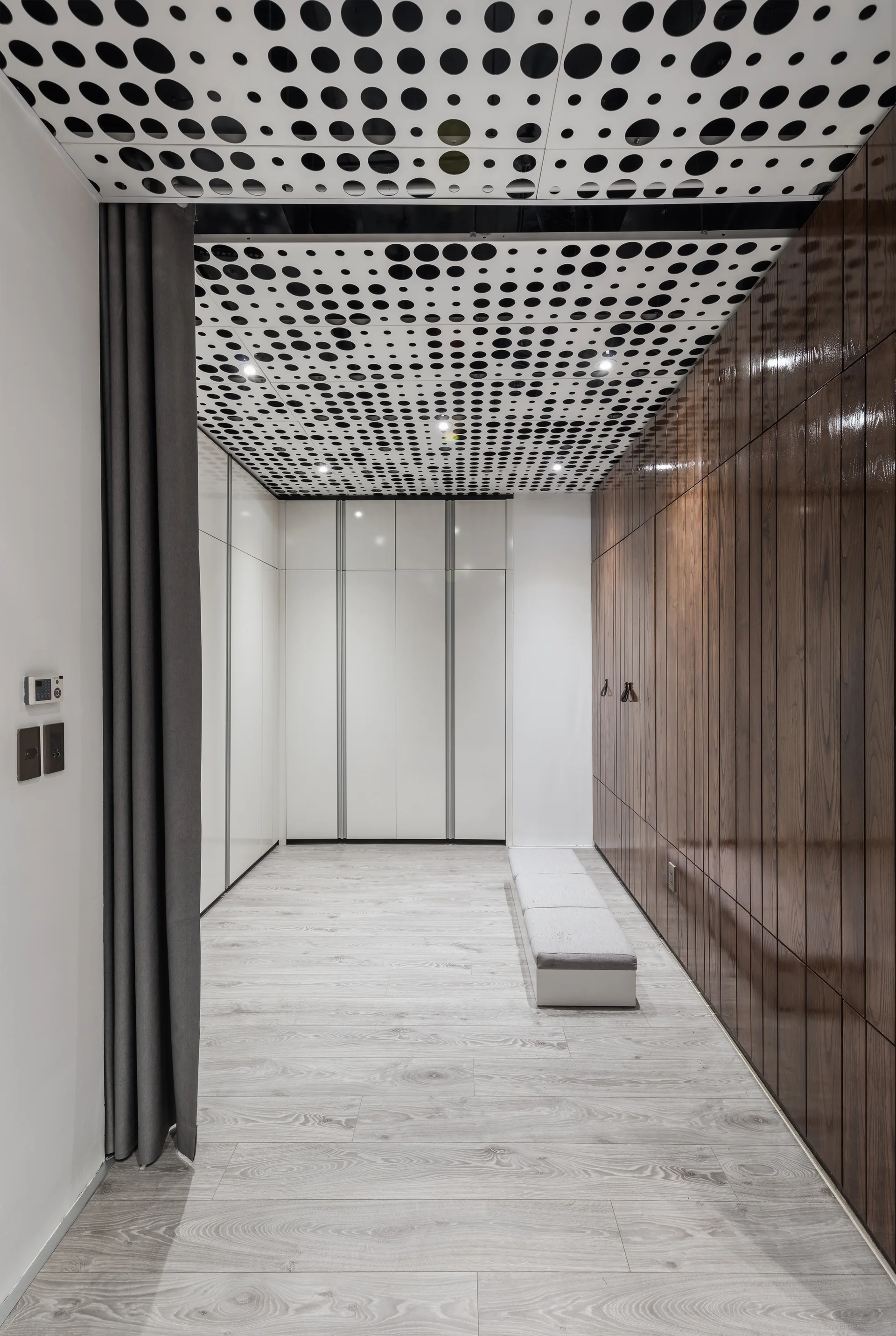 A modern dressing room featuring high-gloss wood and white built-in cabinets, a low bench, grey curtains, and a polka dot ceiling.