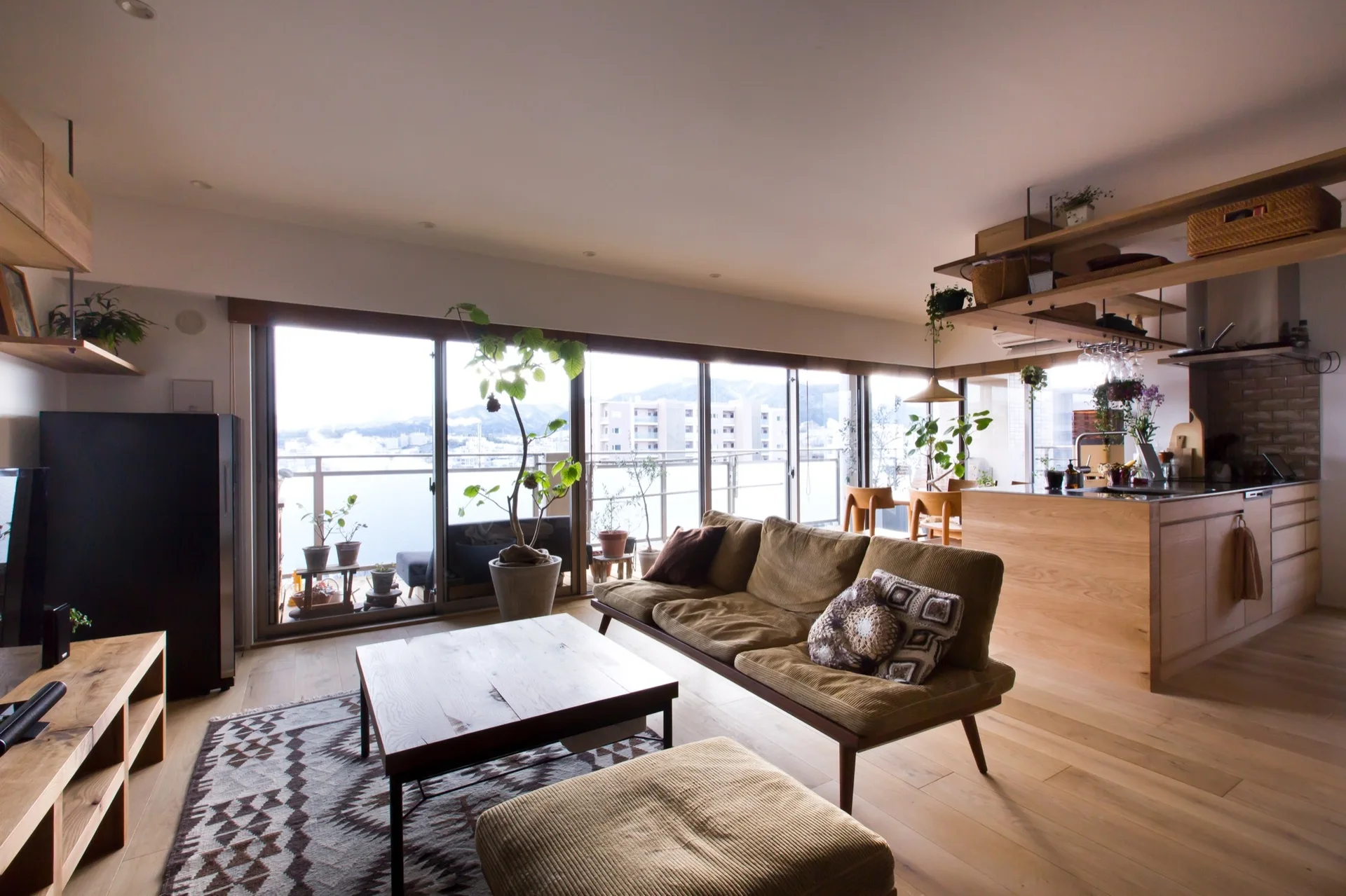 An open-plan apartment living room with a brown corduroy sofa, wooden coffee table, and sliding glass doors leading to a balcony.