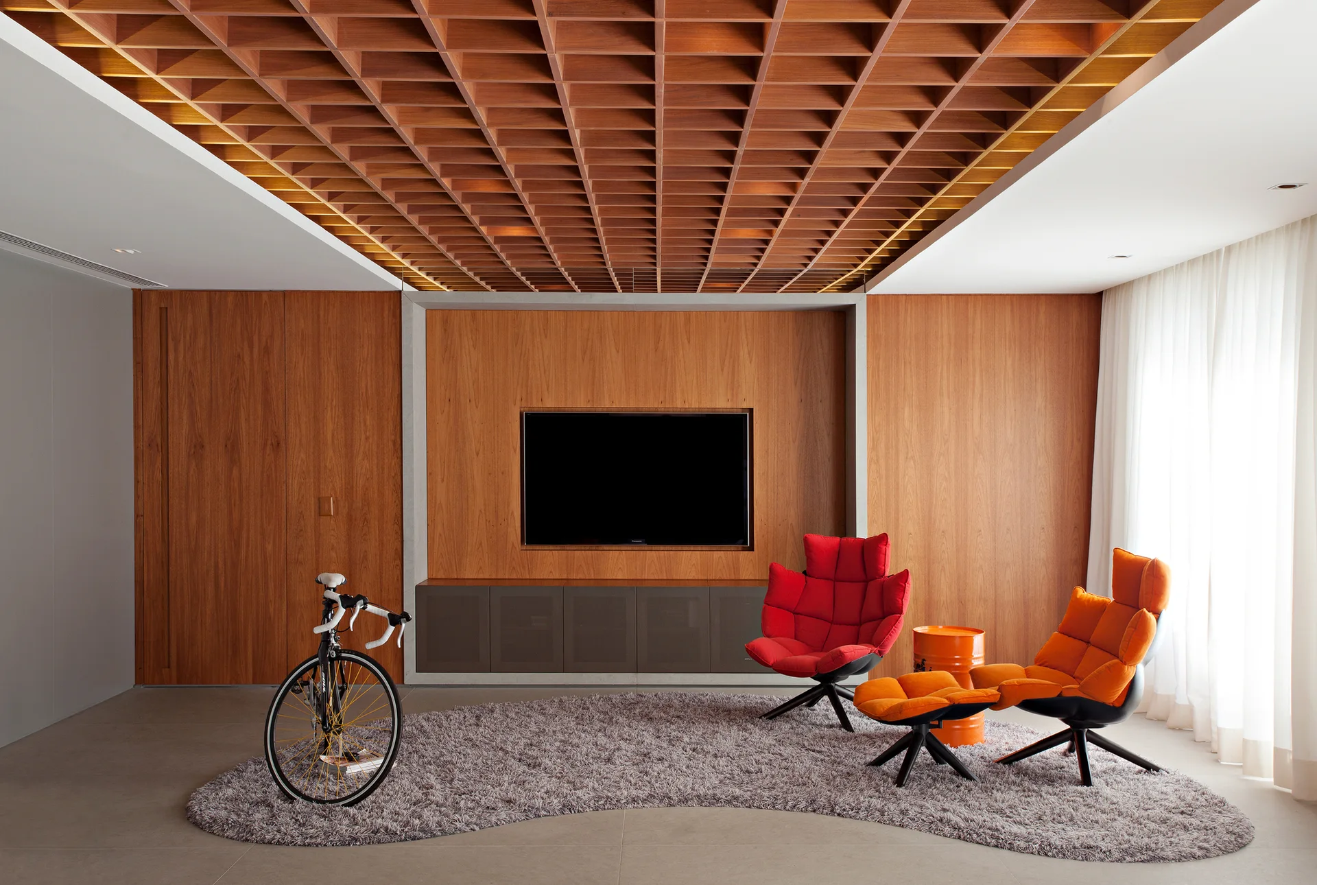 A modern living room features a wooden coffered ceiling, wood-paneled walls, red and orange lounge chairs, and a television mounted in a recessed alcove.