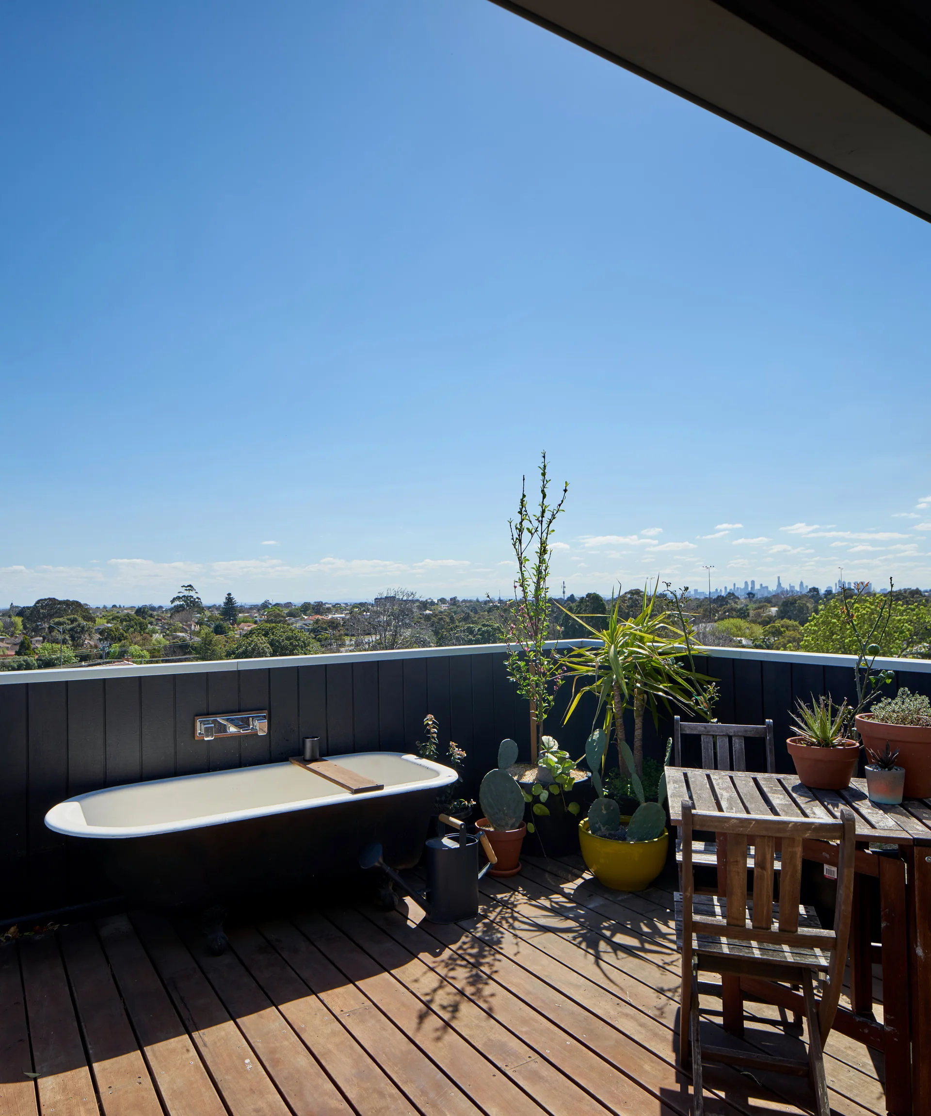 A freestanding bathtub and wooden dining table sit on a rooftop deck surrounded by dark paneling and potted plants.