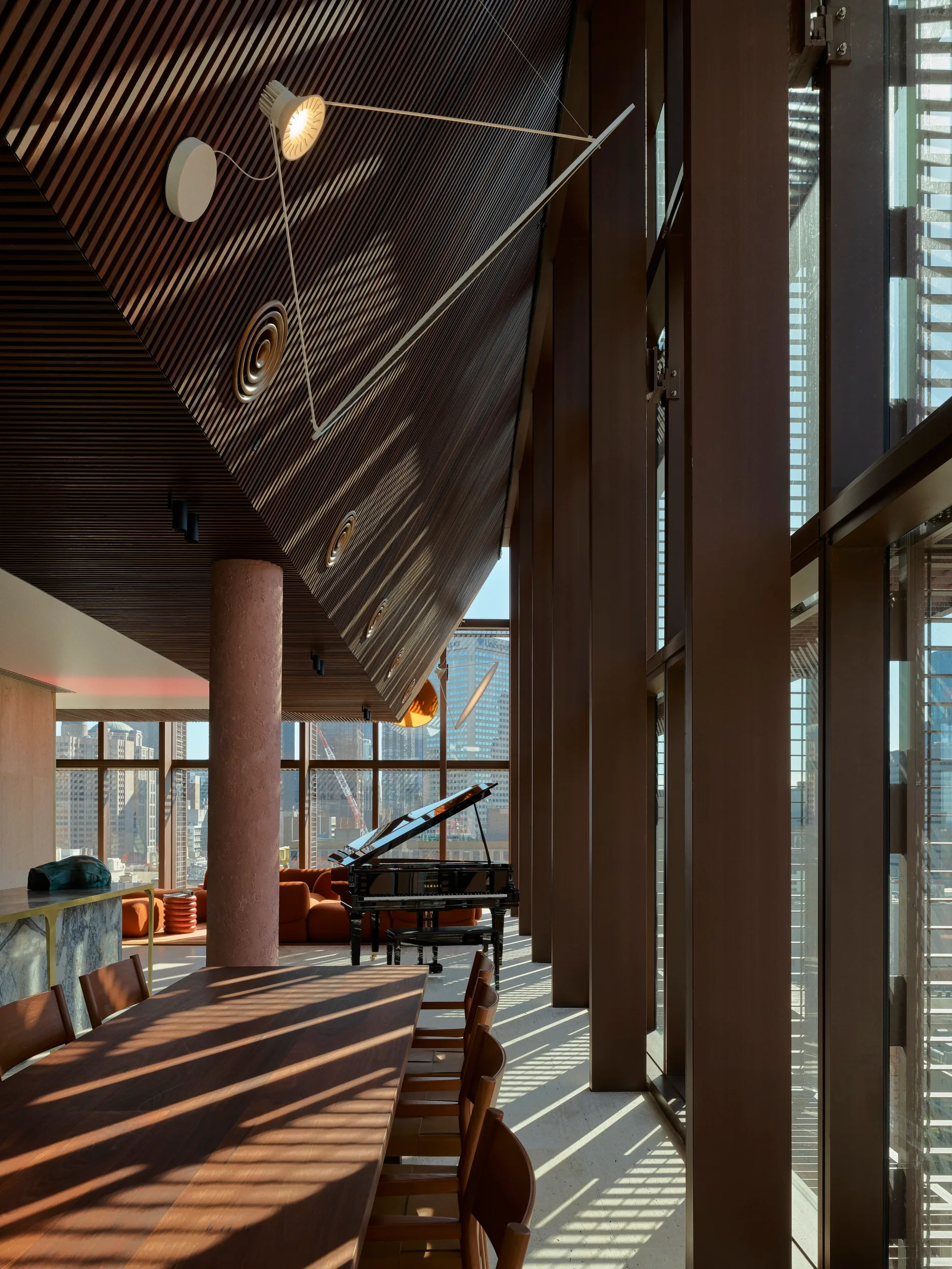 A long wood dining table sits beneath an angled slatted wooden ceiling, next to tall window pillars casting linear shadows on the floor.