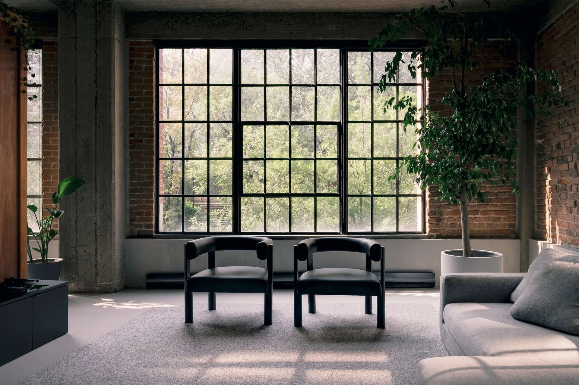 A loft living room features two black armchairs centered in front of a large black grid window, with exposed brick walls and a grey rug.