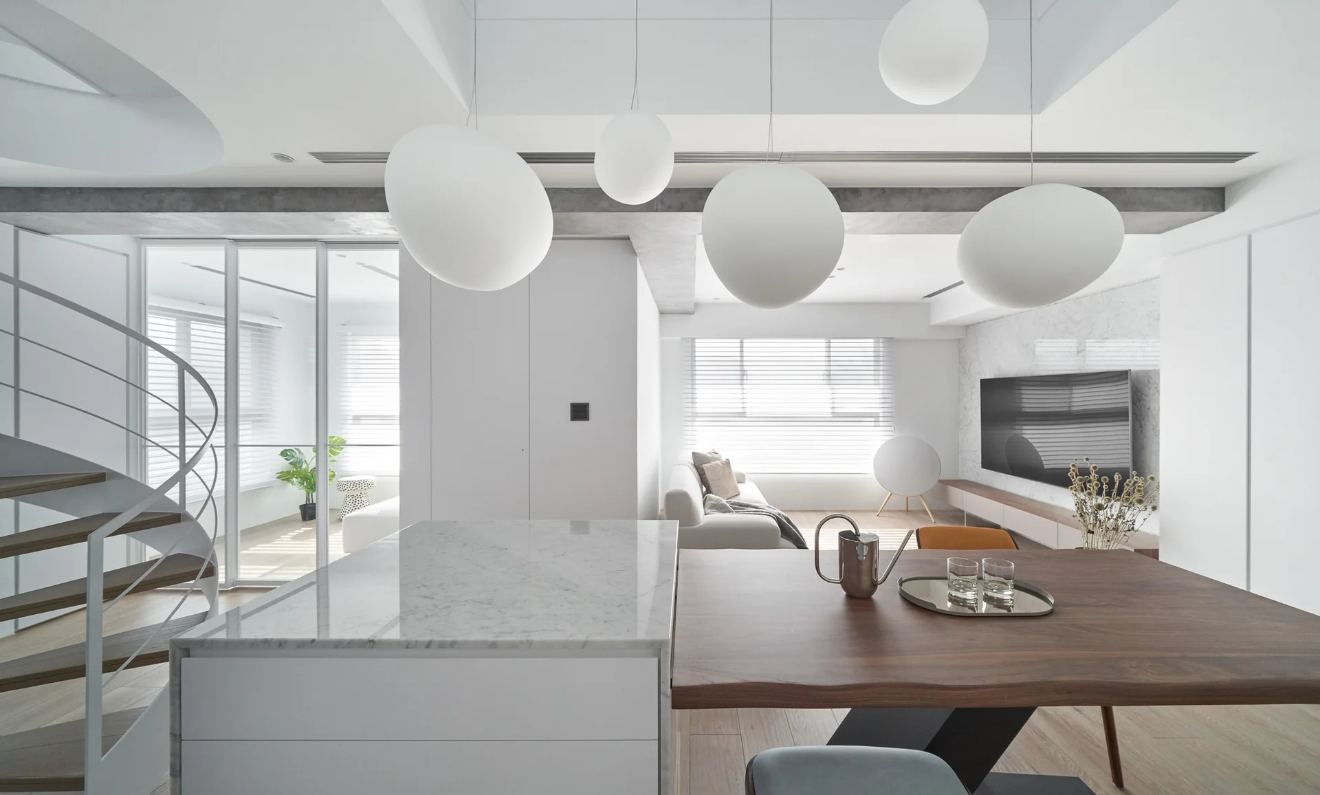 A modern open-plan kitchen and dining area features a marble island connected to a wooden table beneath hanging spherical white pendant lights.