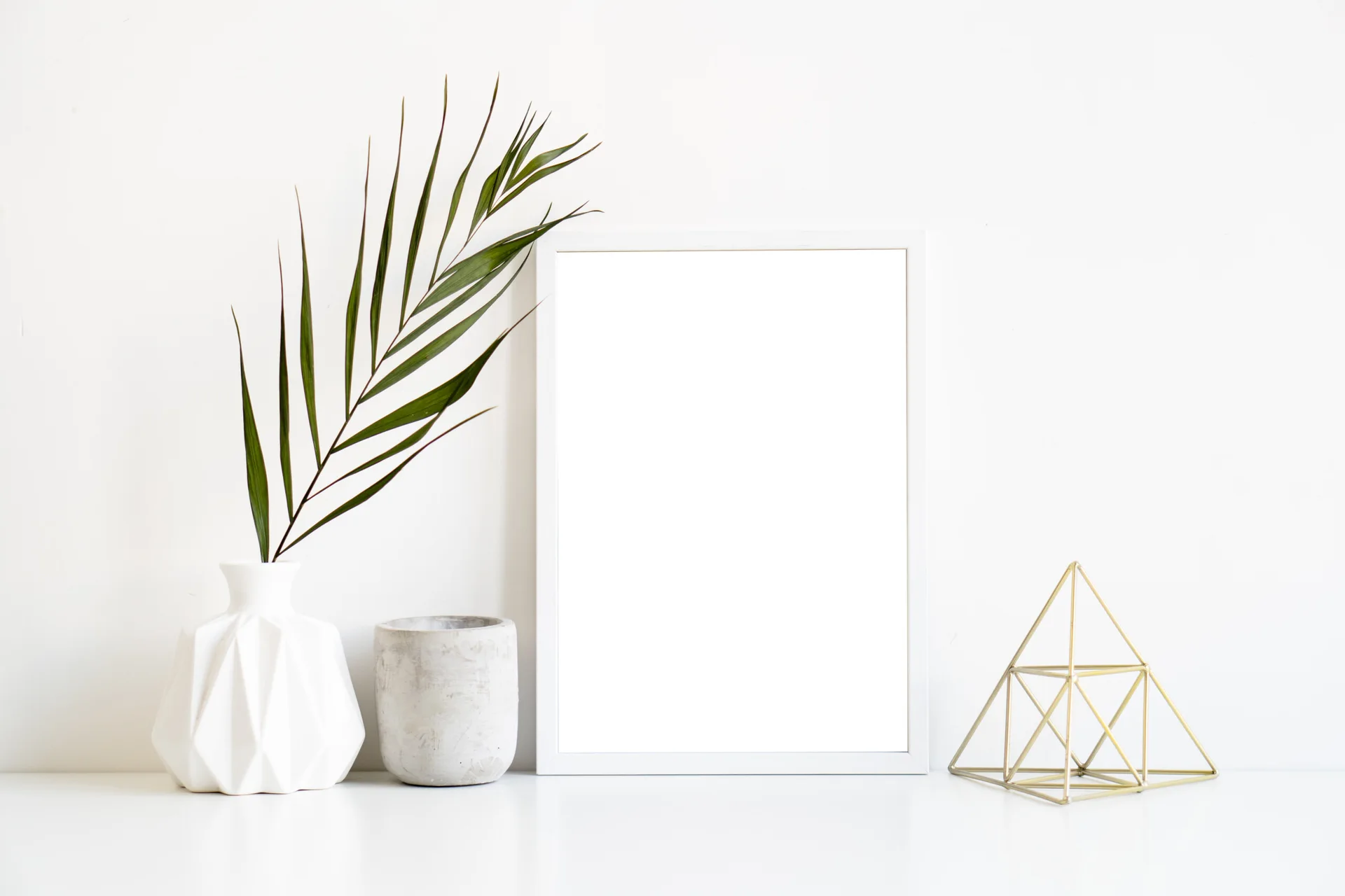 A blank white picture frame sits on a white tabletop alongside a faceted ceramic vase with a palm leaf, concrete planter, and gold wire pyramid.