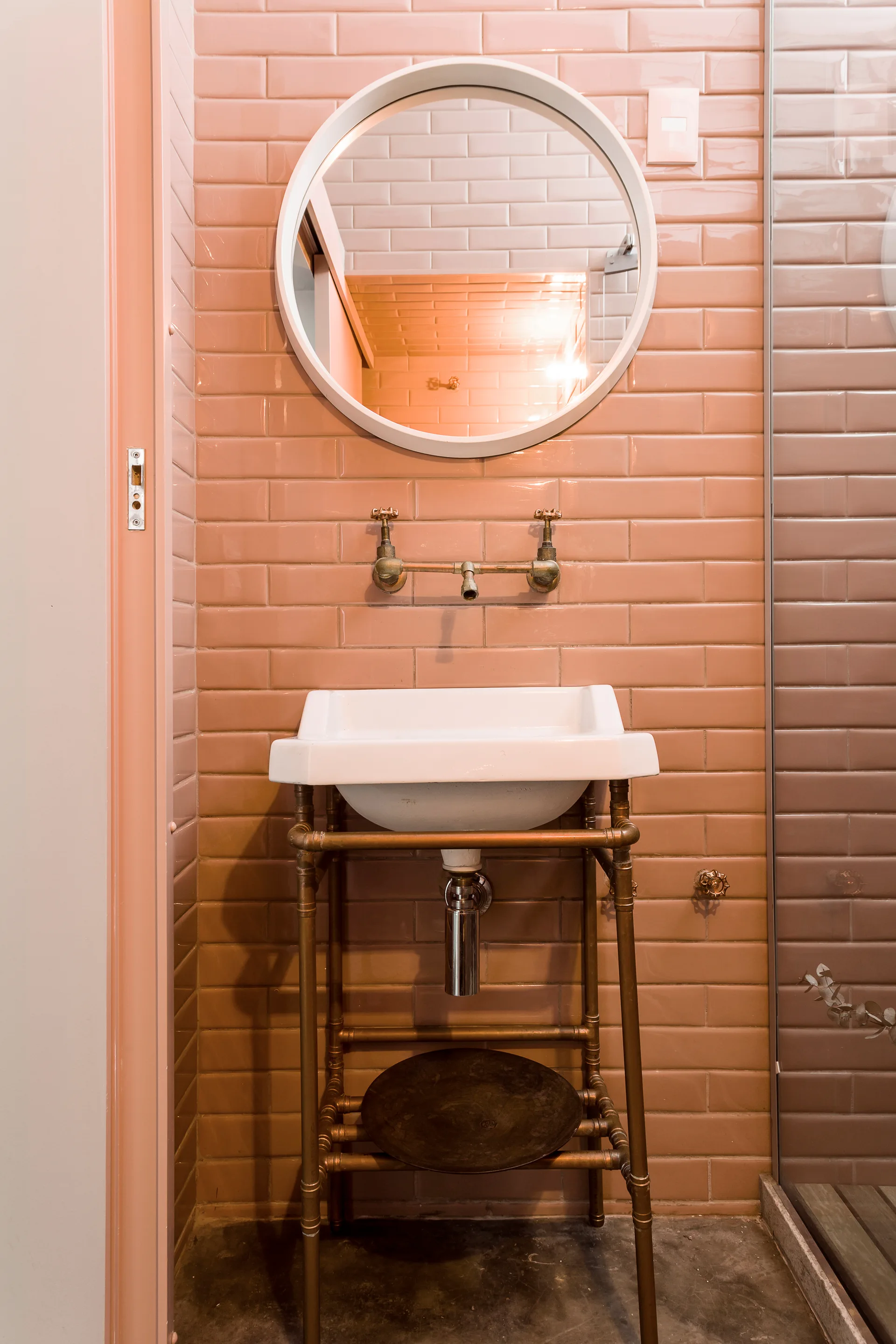 A white basin sink on an industrial brass stand is set against pink subway tiles beneath a circular white-framed mirror.